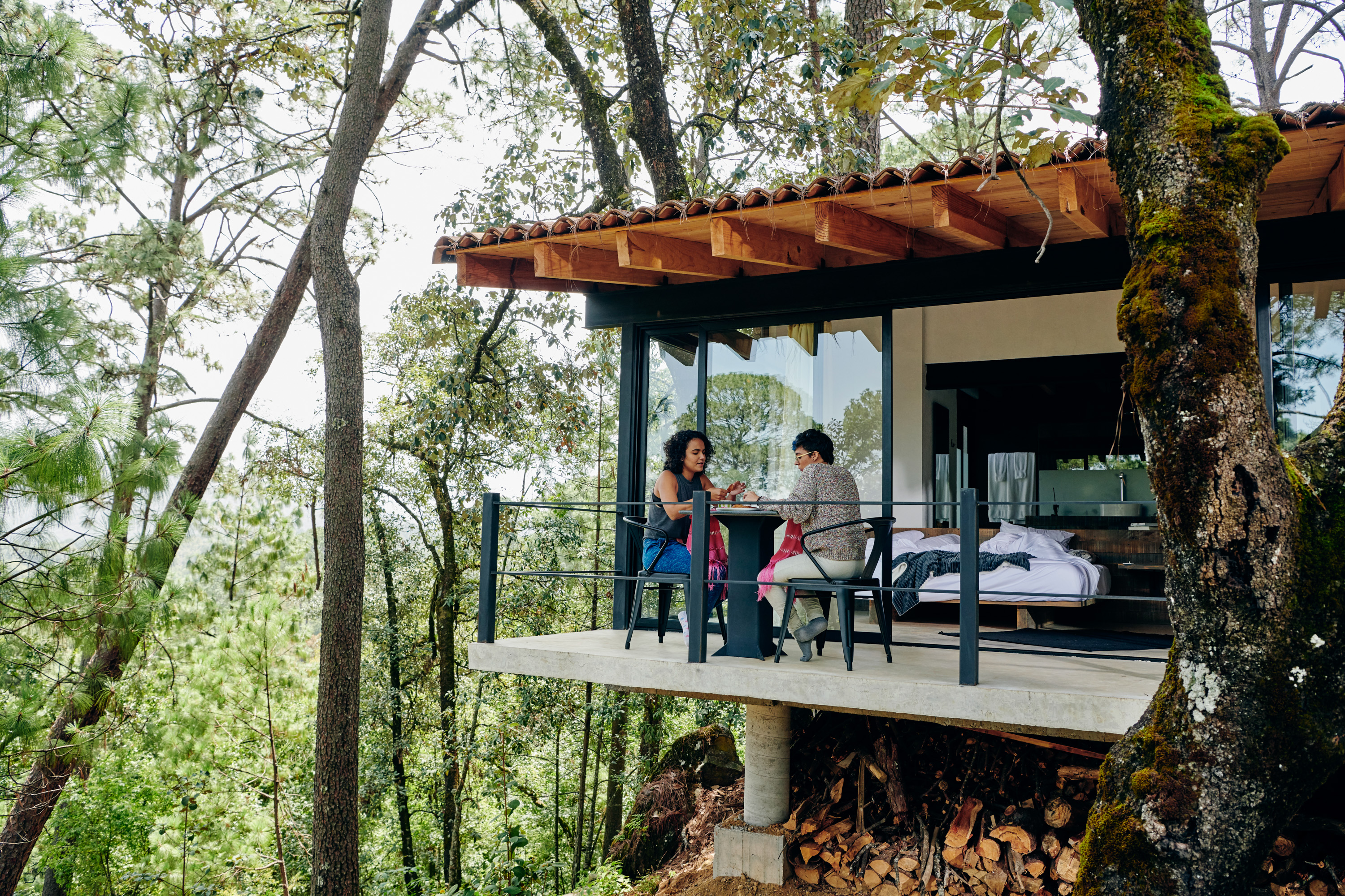 Two travel companions sit at a small table on the balcony of a forest treehouse, surrounded by tall, green trees.