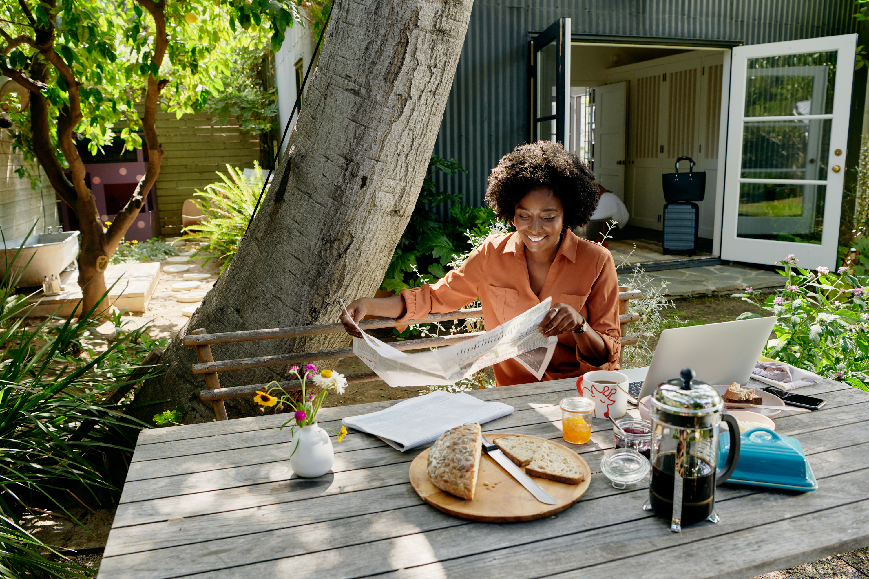 Woman dressed in an orange shirt is seated at a patio table outside her listing reading the newspaper, eating breakfast and working on her laptop.