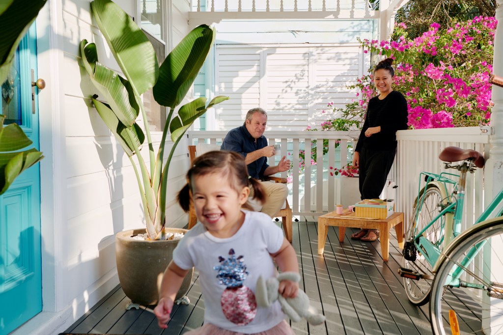 Toddler girl in foreground running towards camera on a front porch of an Airbnb. Her parents dressed in black casual clothes are watching over her in the background while enjoying their morning coffee.