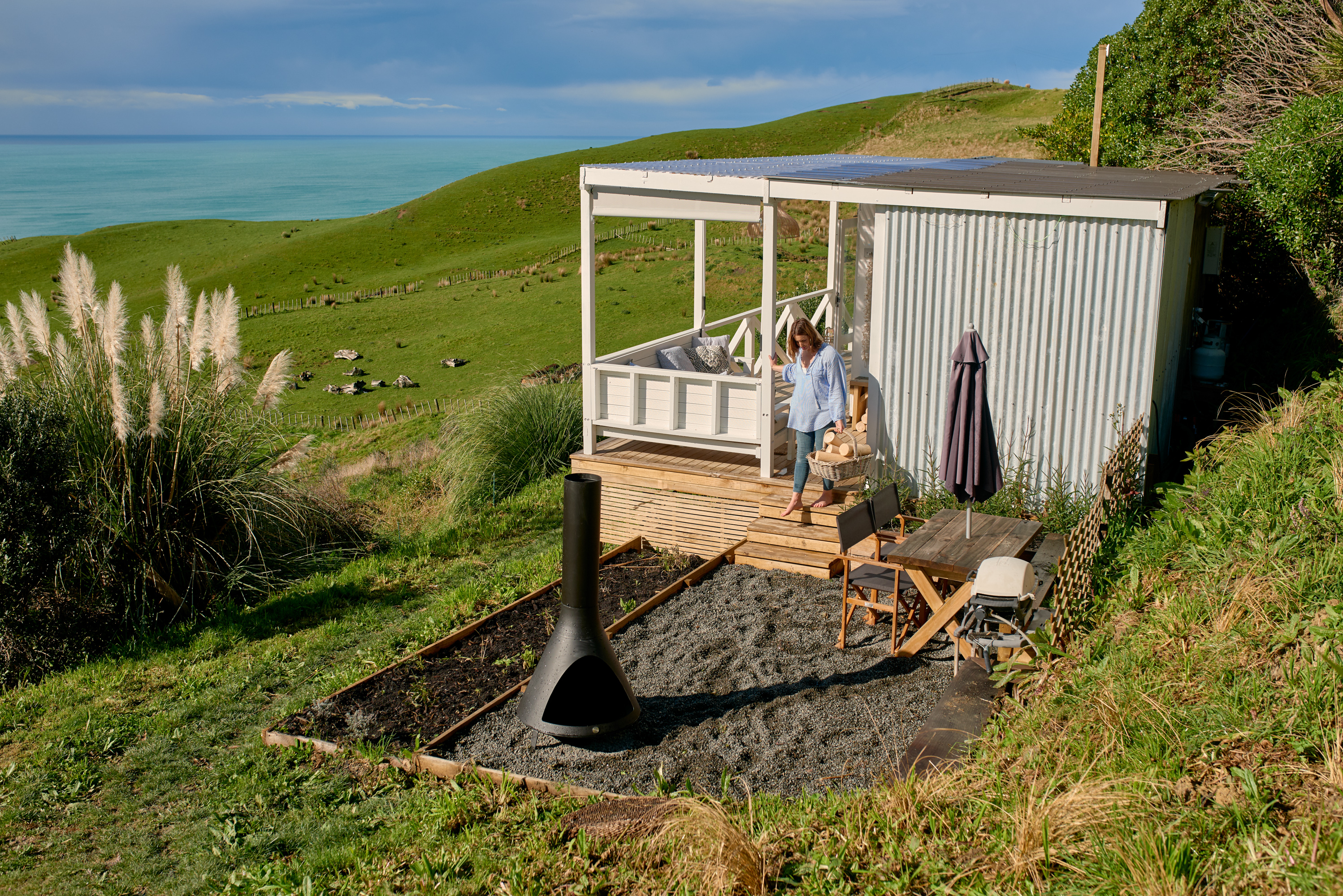 Woman in a striped button down shirt descends down the side steps of a renovated shipping container home with basket in hand. She's stepping out onto a gravel side yard with a fire pit and the home is situated on a grassy bluff overlooking the coastline.