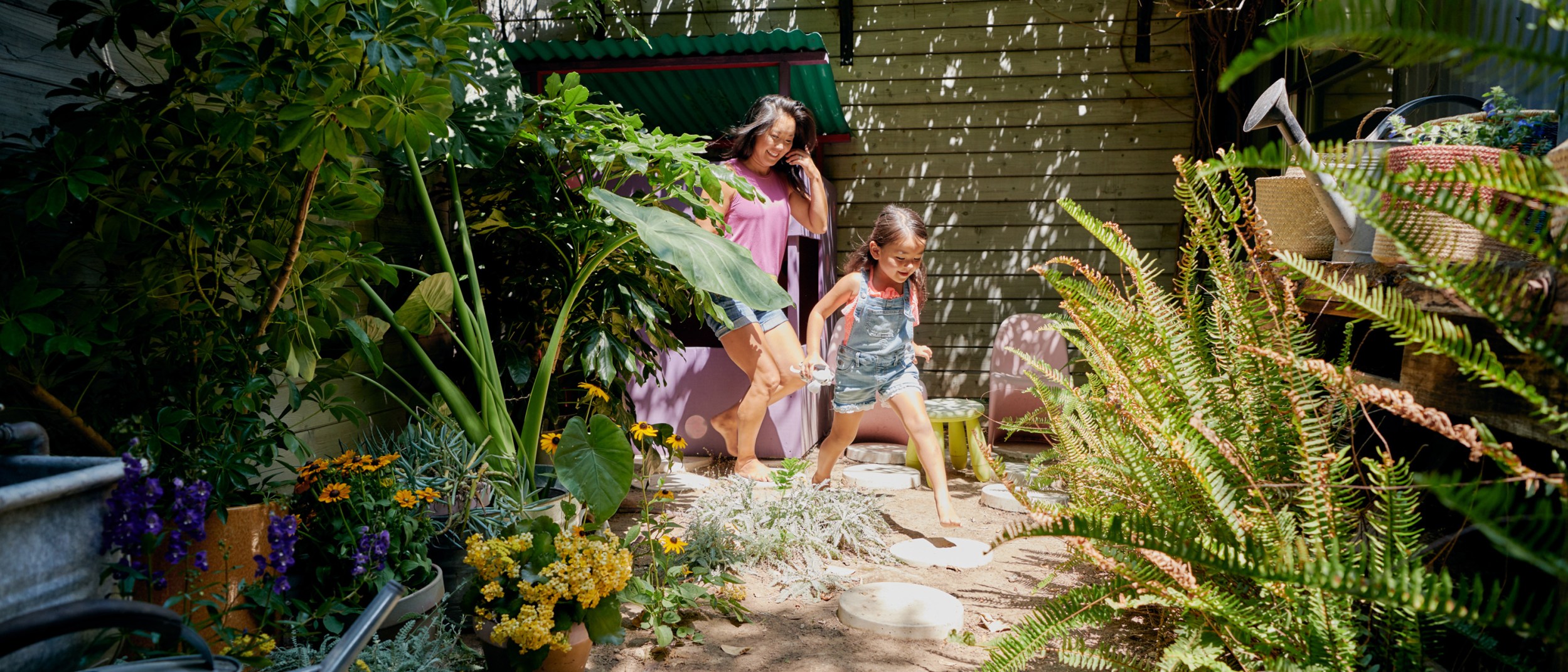 Mother and daughter running through a verdant backyard patio surrounded by flowers and greenery.