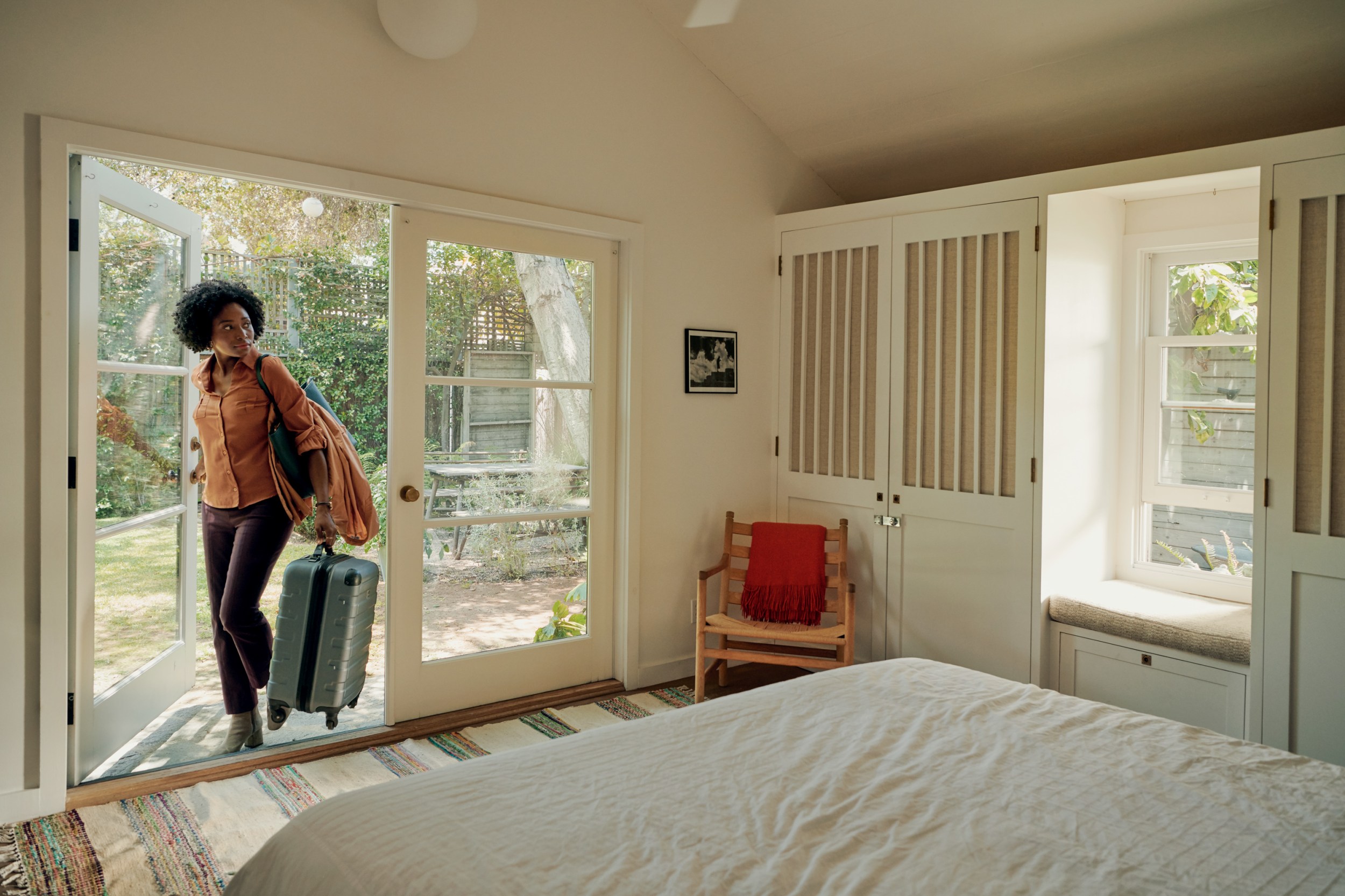 Woman carrying a suitcase and purse entering a sunny white bedroom