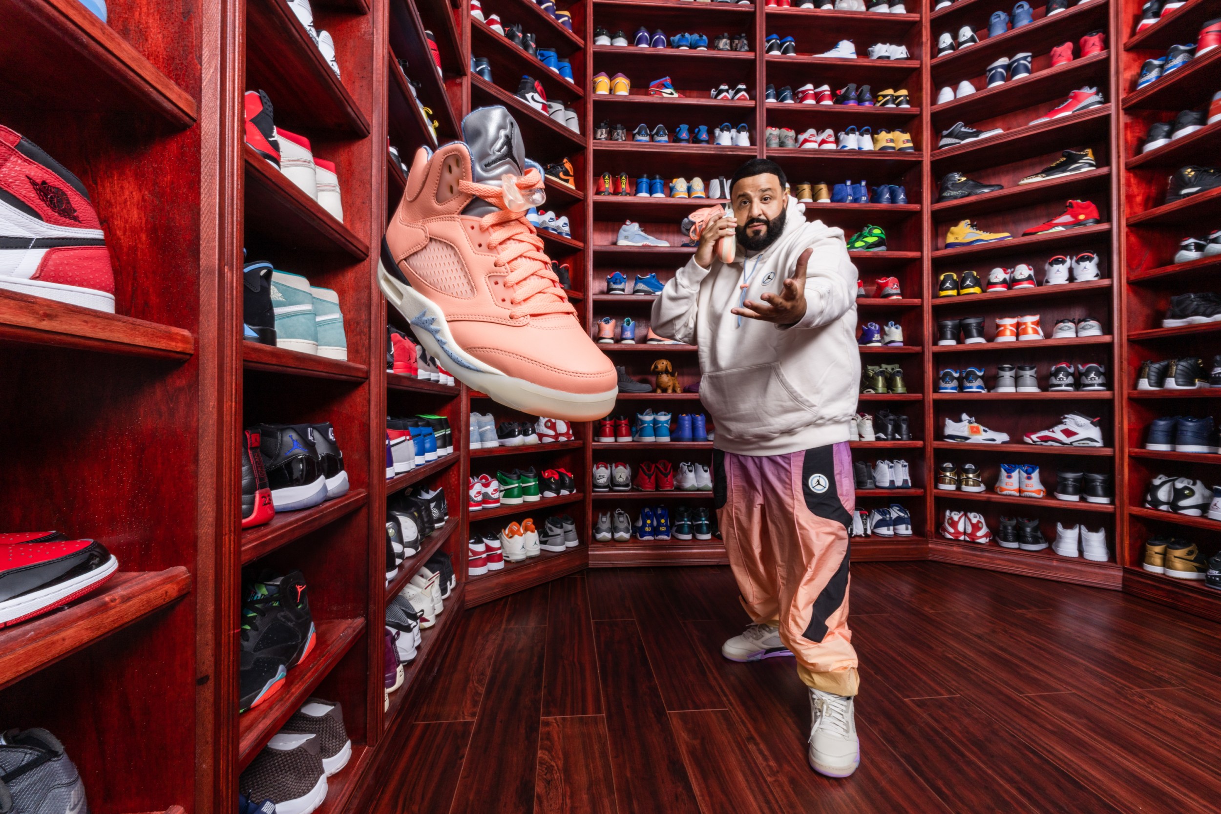 DJ Khaled stands in the center of a large shoe closet surrounded by shelves of bright colored sneakers. He holds a pink one up to his ear like a telephone, with another floating in the air as if he tossed it to the audience.