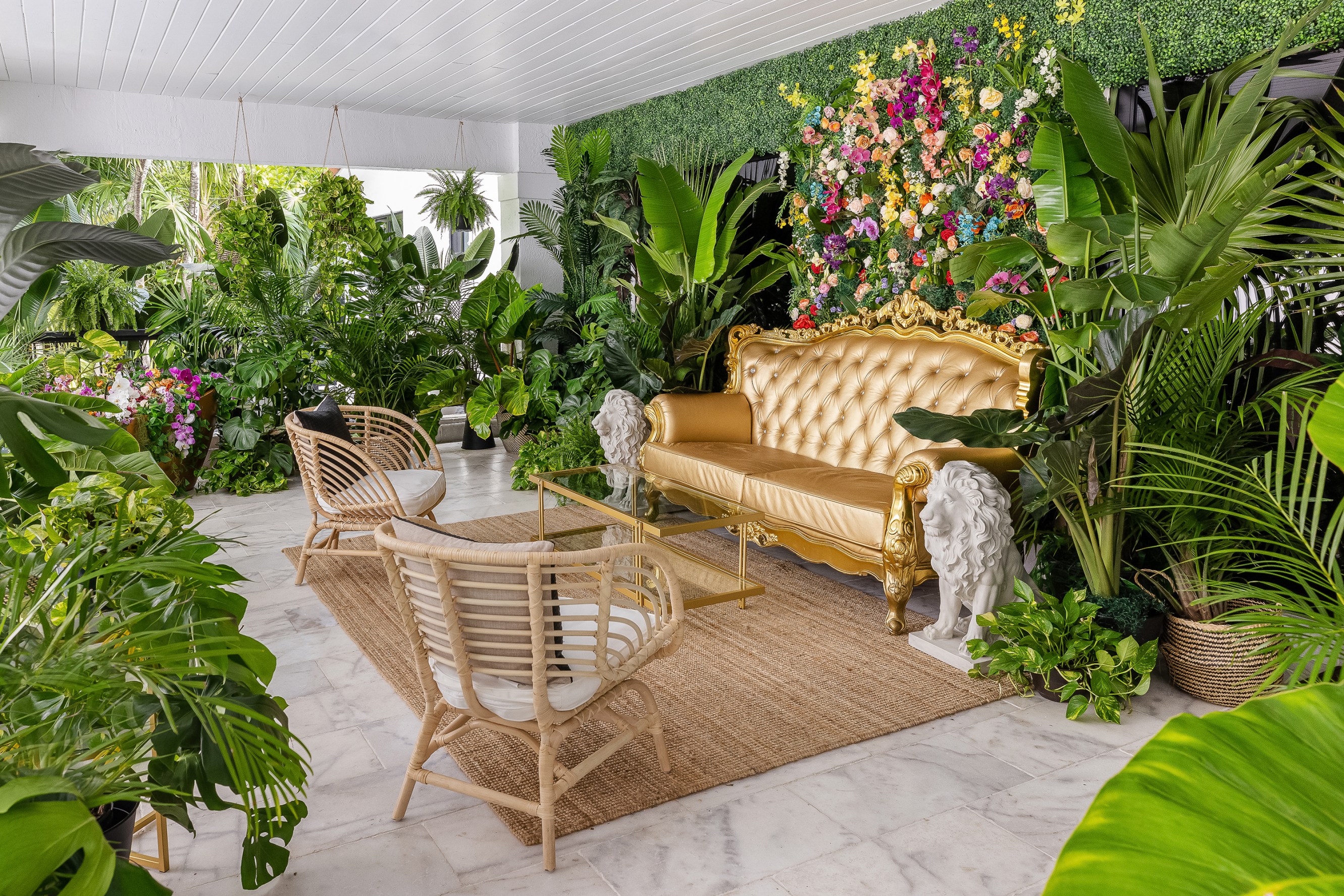 An outdoor seating vignette showing a gold plush sofa and two arm chairs facing it, with a floral wall backdrop surrounded by lush green plants.