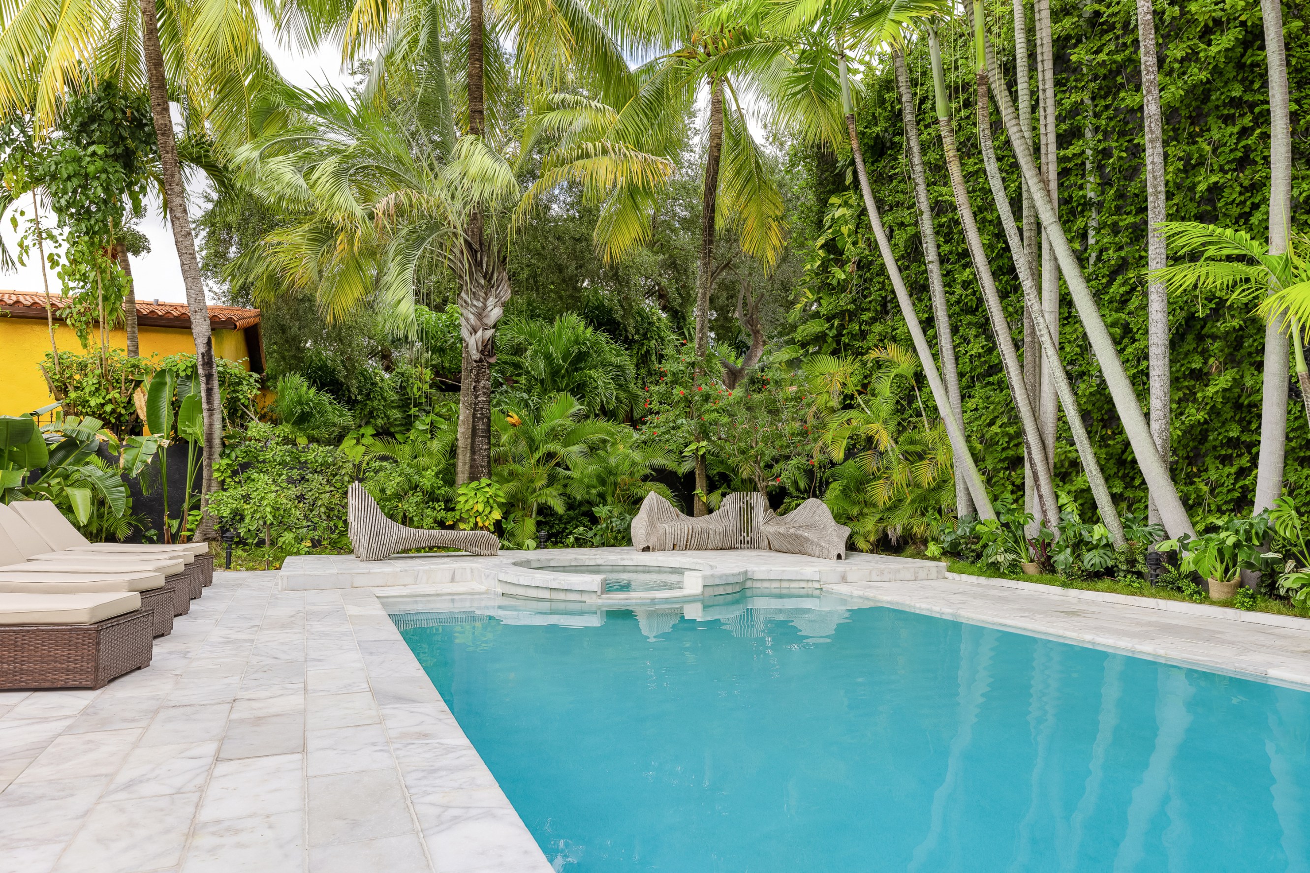 Backyard pool in the foreground with a couple of chaise lounge chairs and artistic wooden sofas on the far side of the pool. Palm trees and other lush greenery surround in the background.