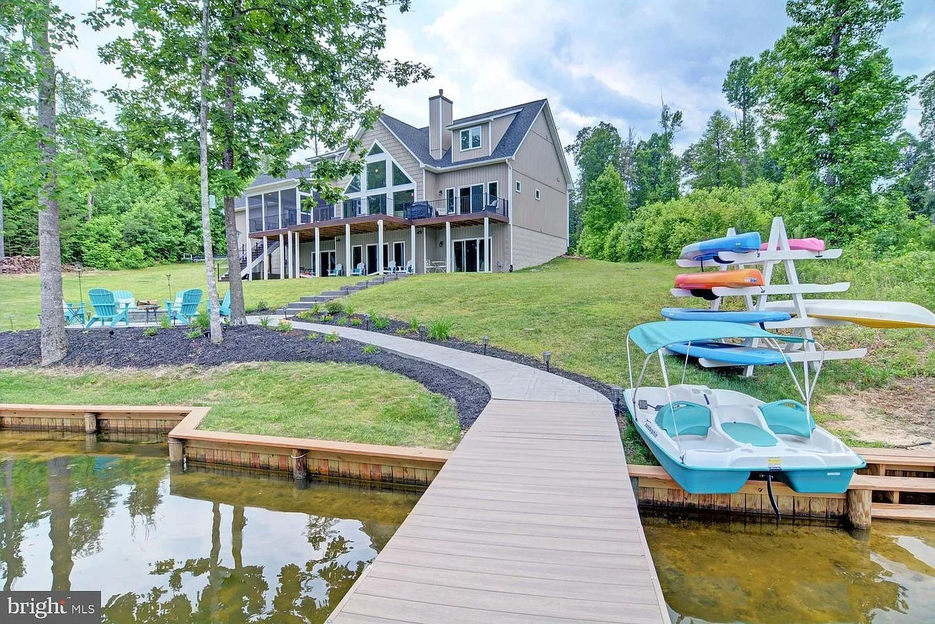 Two-story, house with floor to ceiling back windows, and a path leading to a dock along a lake with a paddle boat docked nearby