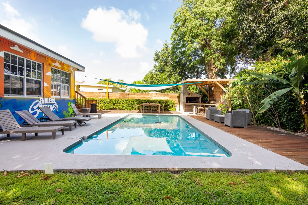 A brown one-story house with big glass windows next to a pool with three pool chairs, next to trees