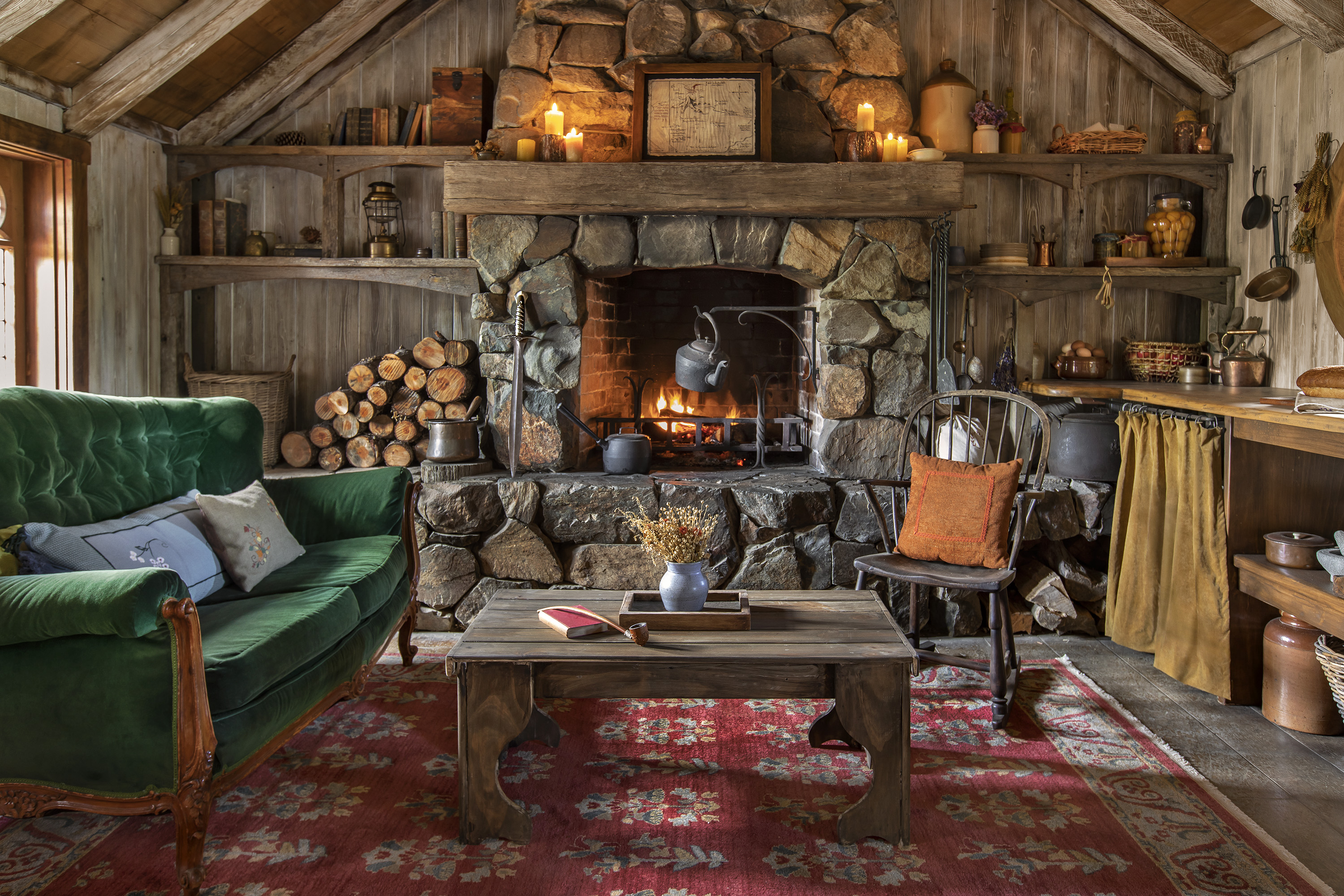 Living room view showing a plush green couch, a red carpet and wooden coffee table with a vase of dried flowers on top. Behind the seating area is a roaring fireplace with a hot tea kettle suspended above it and firewood alongside the stone hearth. A wooden mantel is nestled above it with lit pillar candles and what appears to be a framed map.e