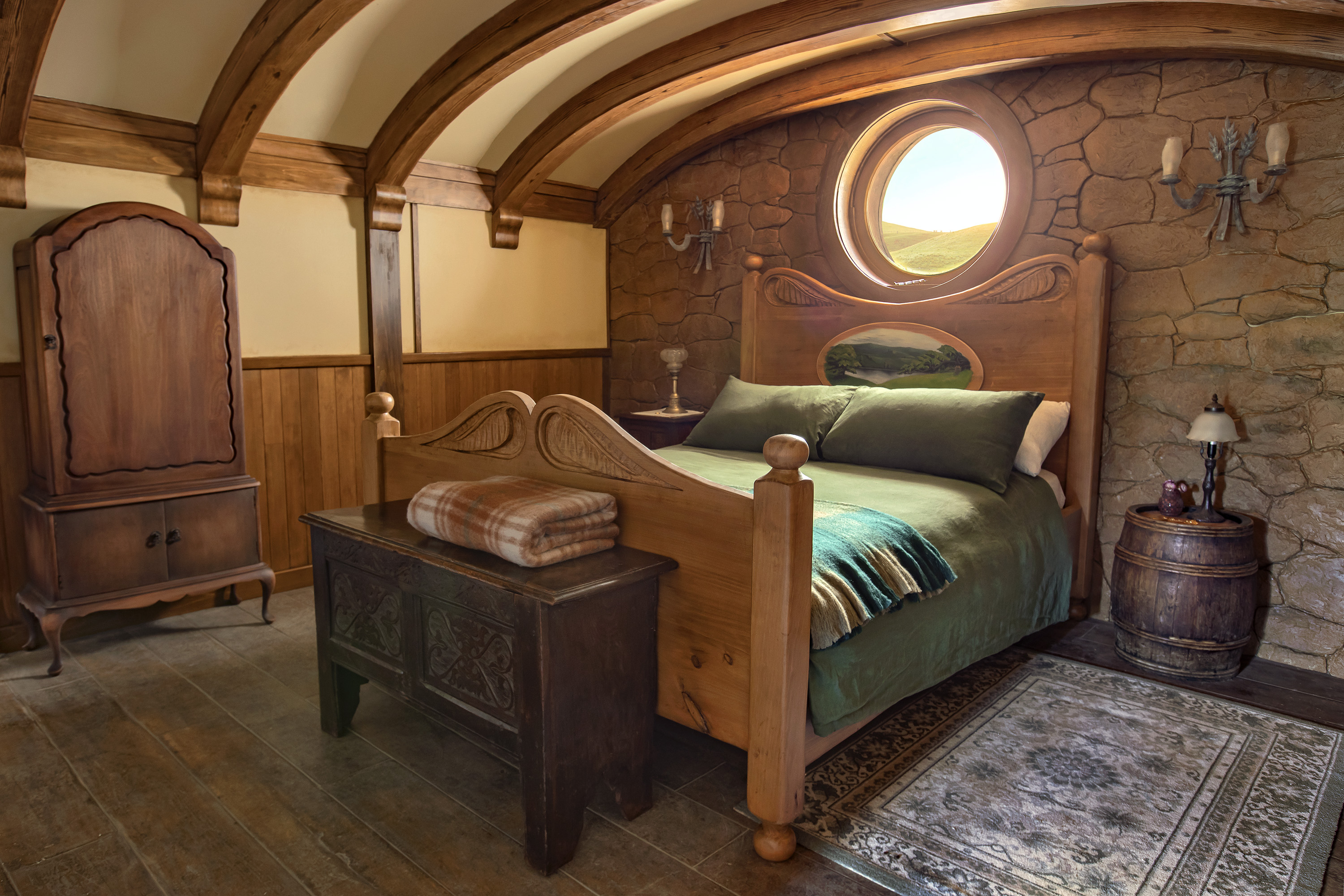Bedroom view with wood floors, a wooden chest closet and a large wooden bed with green pillows and blankets. A small oriental rug lays alongside the bed and a wooden storage bench is at the end of the bed. A small porthole window is above the bed's headboard.