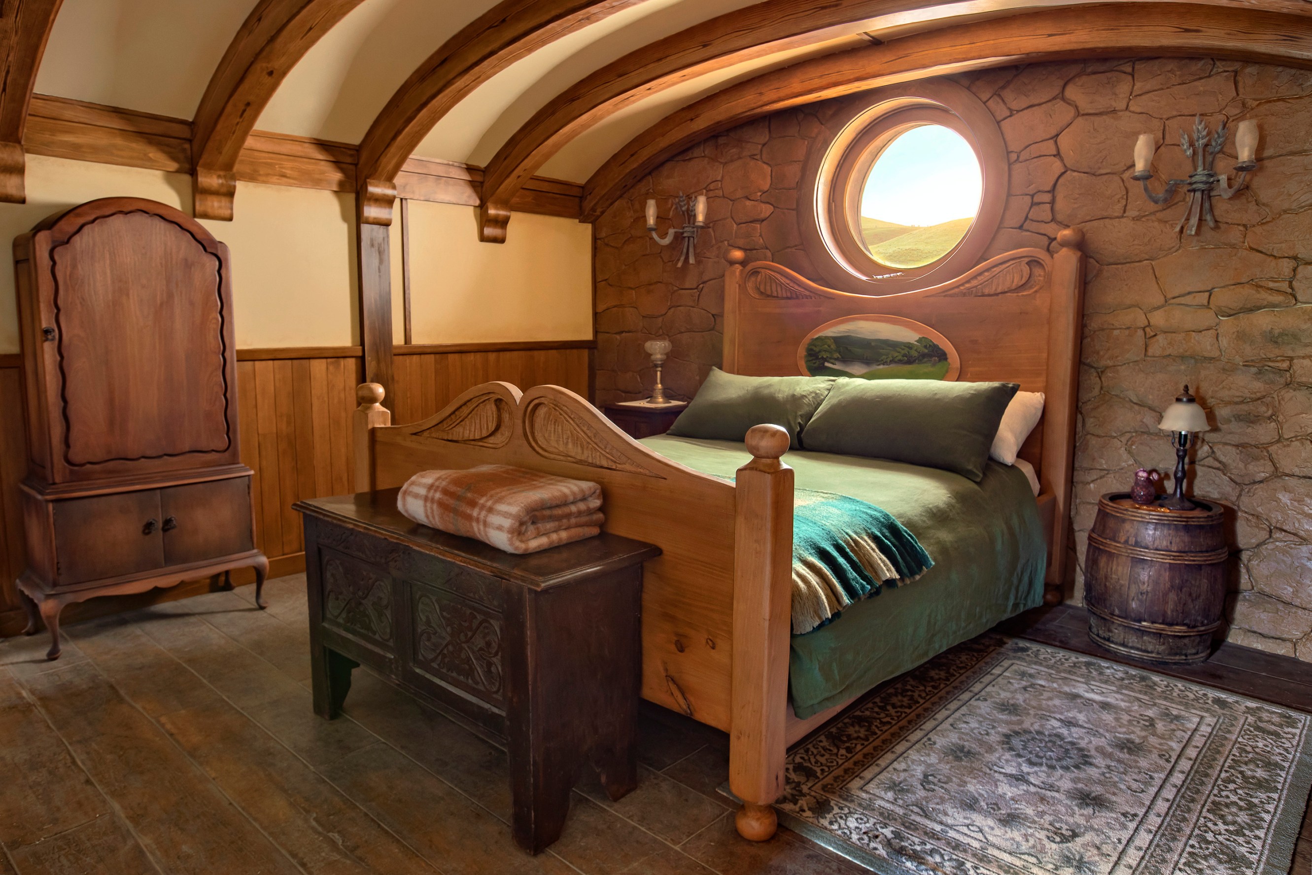 Bedroom view with wood floors, a wooden chest closet and a large wooden bed with green pillows and blankets. A small oriental rug lays alongside the bed and a wooden storage bench is at the end of the bed. A small porthole window is above the bed's headboard.