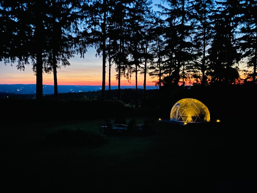 A golden orb artificial igloo with a tree line and sunset view behind