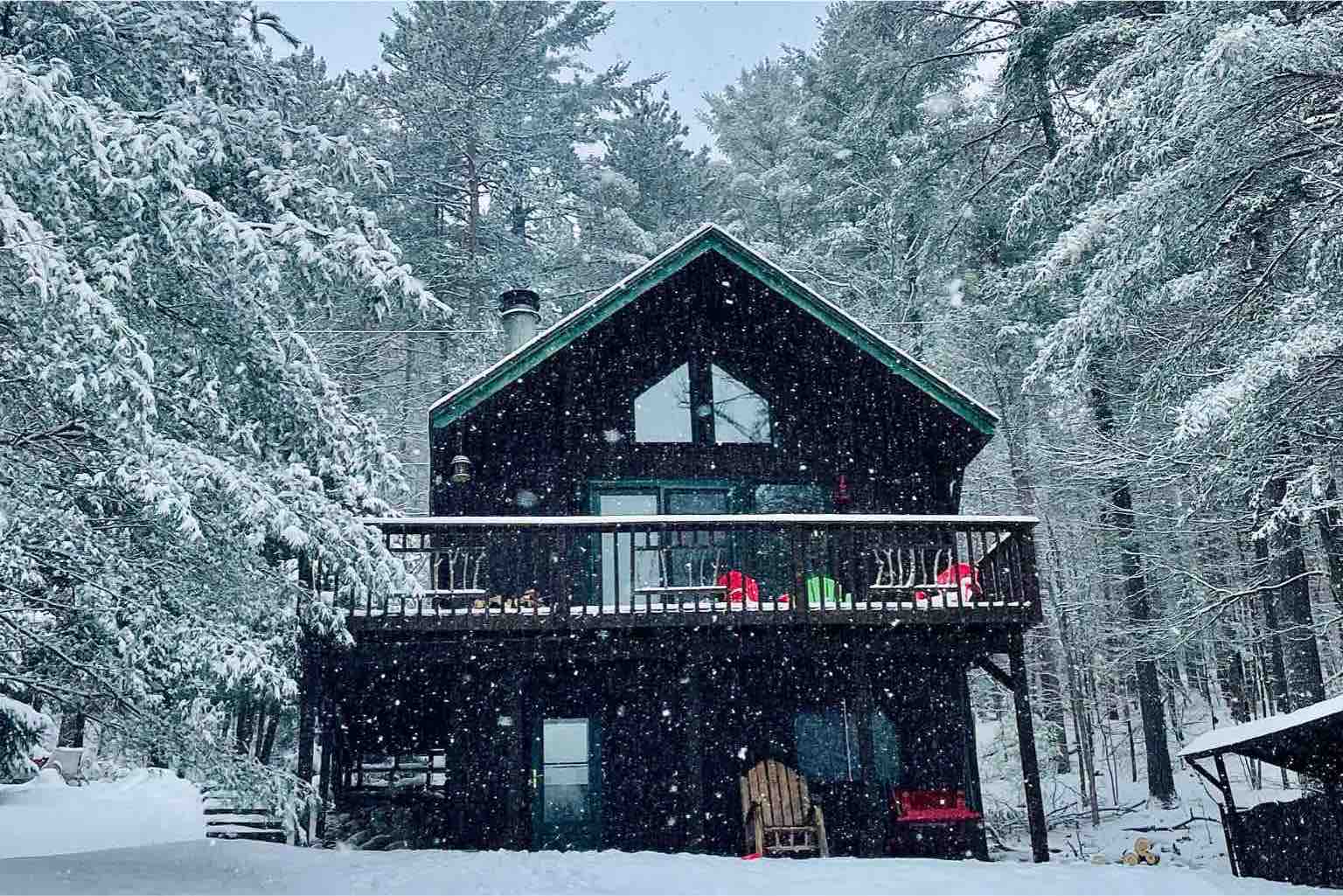 Two story brown cabin with a wrap around deck surrounded by snow-covered tree and snow falling and covering the ground