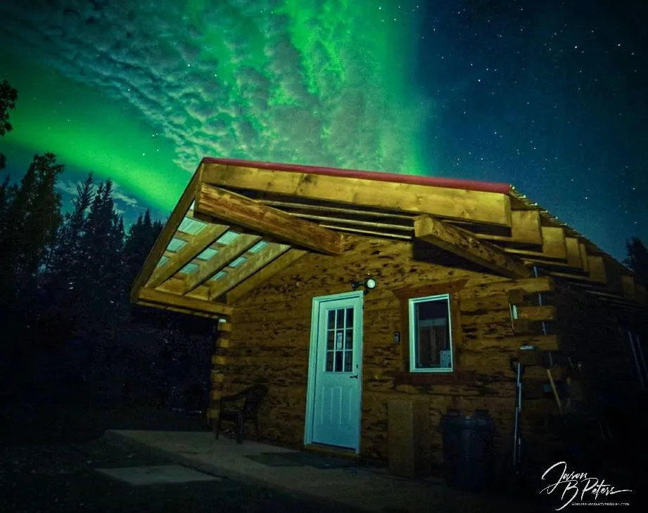 A wooden cabin with a white door set against the backdrop of vibrant green Northern Lights aurora in the night sky