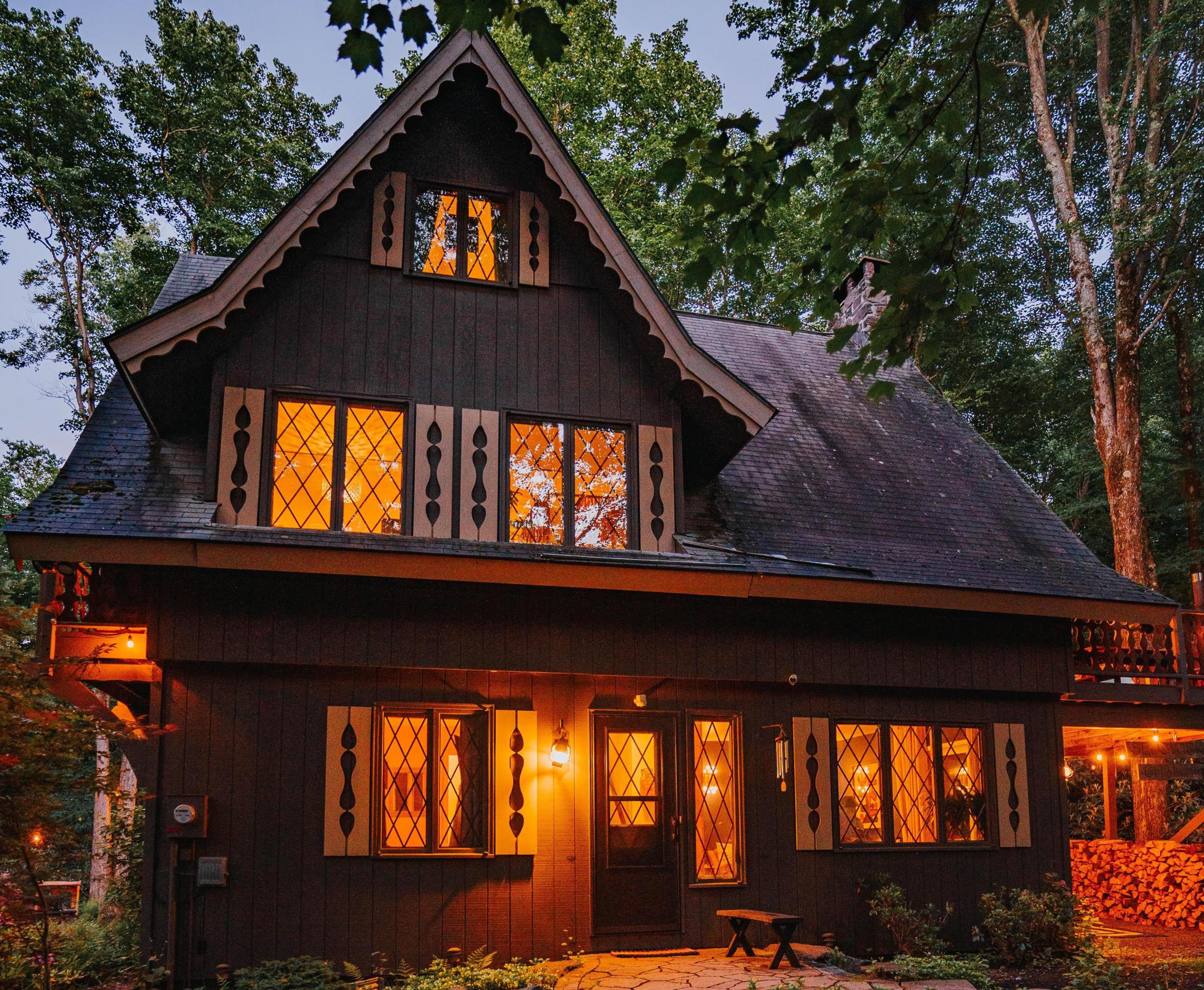 Two-story cabin with an A-frame second story, with warm yellow lighting, surrounded by trees