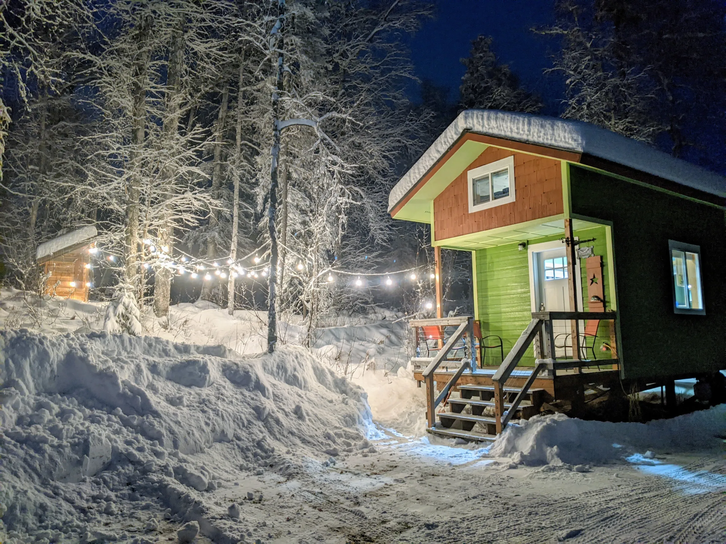 Well-light two-story, tiny home cabin with a wooden step porch; the lower level is green and the house is surrounded by snowy ground and bright festive lights strung in the trees