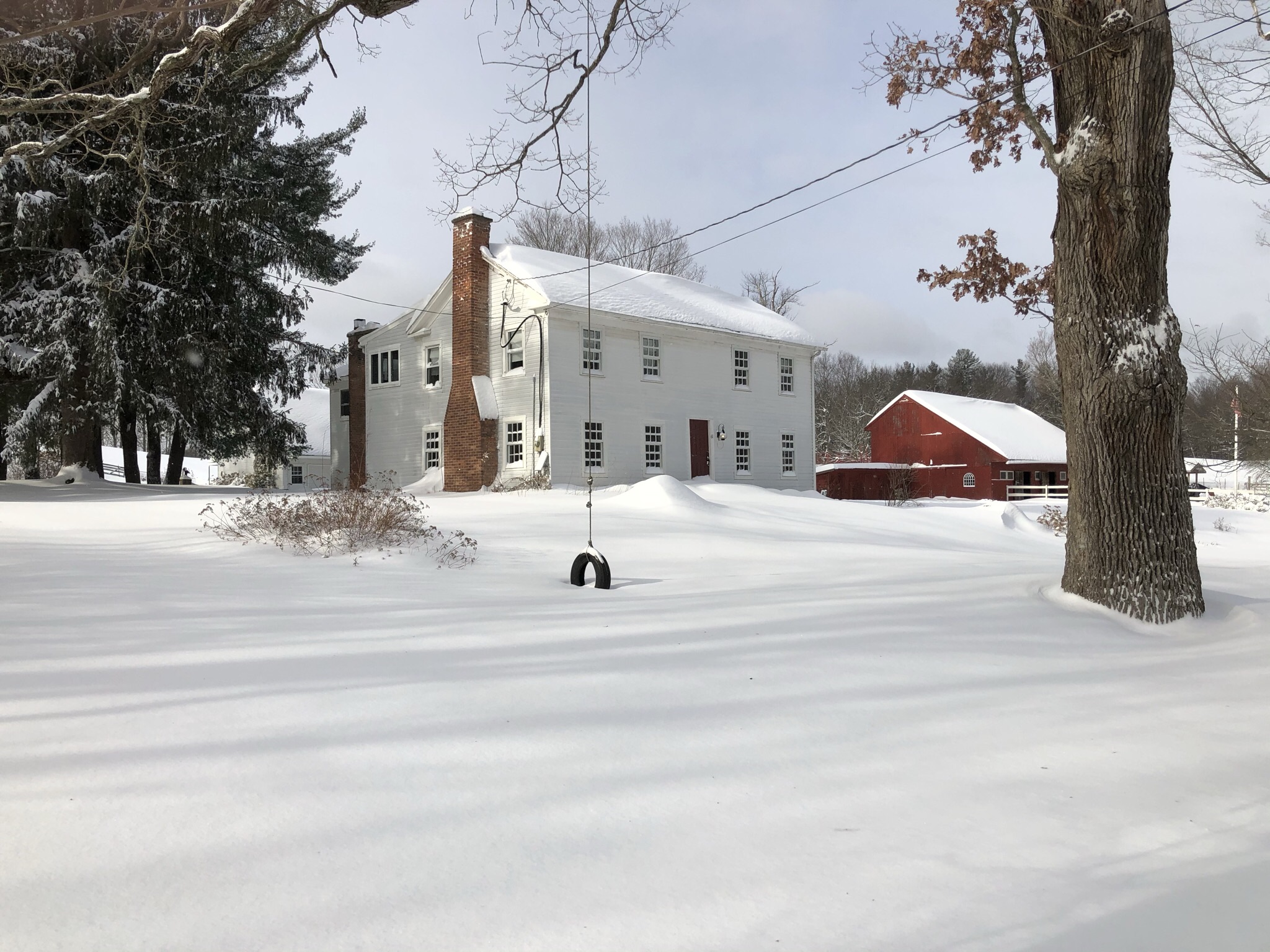 White two story home next to a red barn in front of a snowy yard with a tree and tire swing