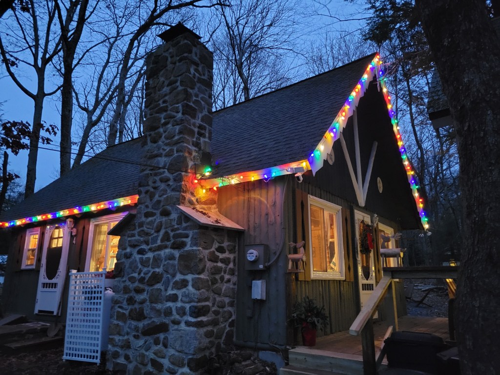 Gingerbread cabin with a brick chimney on one side; the house is strung with Christmas lights