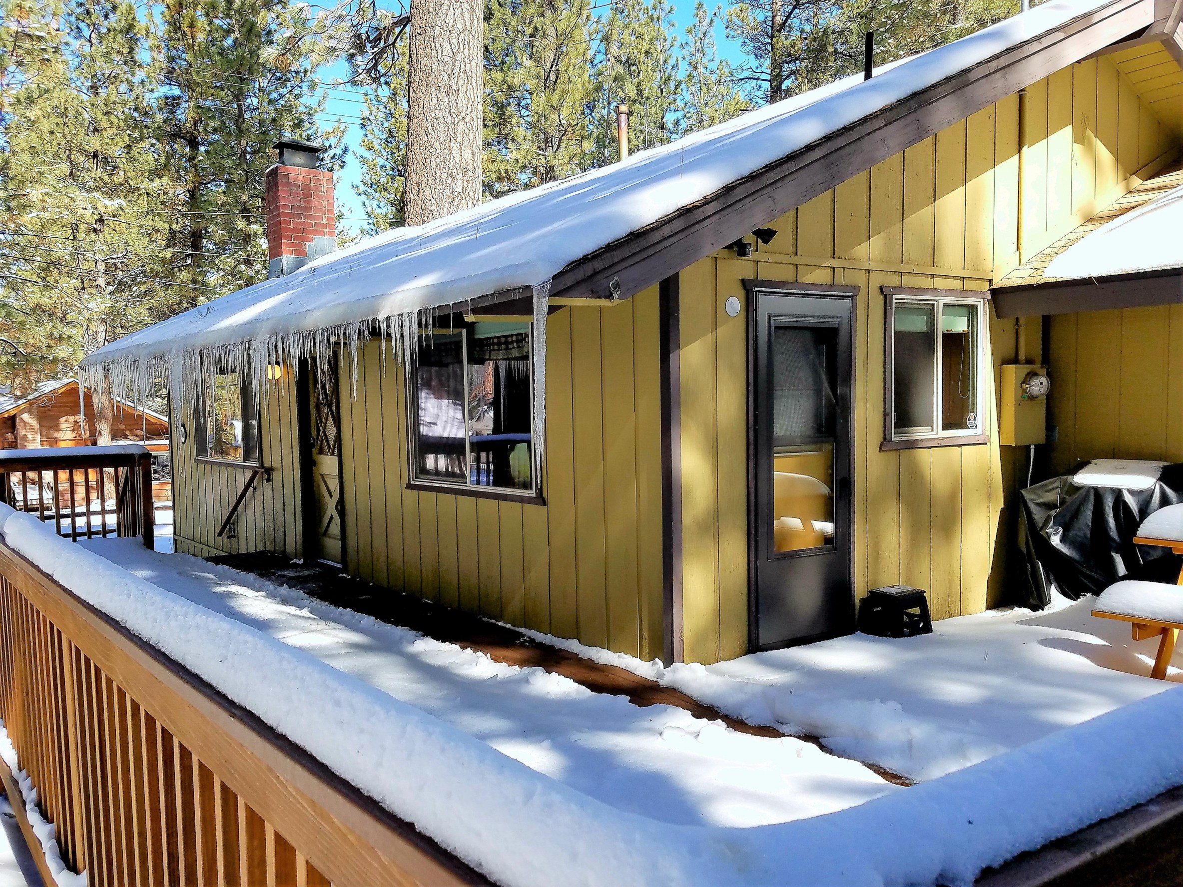 A small yellow cabin with a snow covered roof and porch surrounded by trees