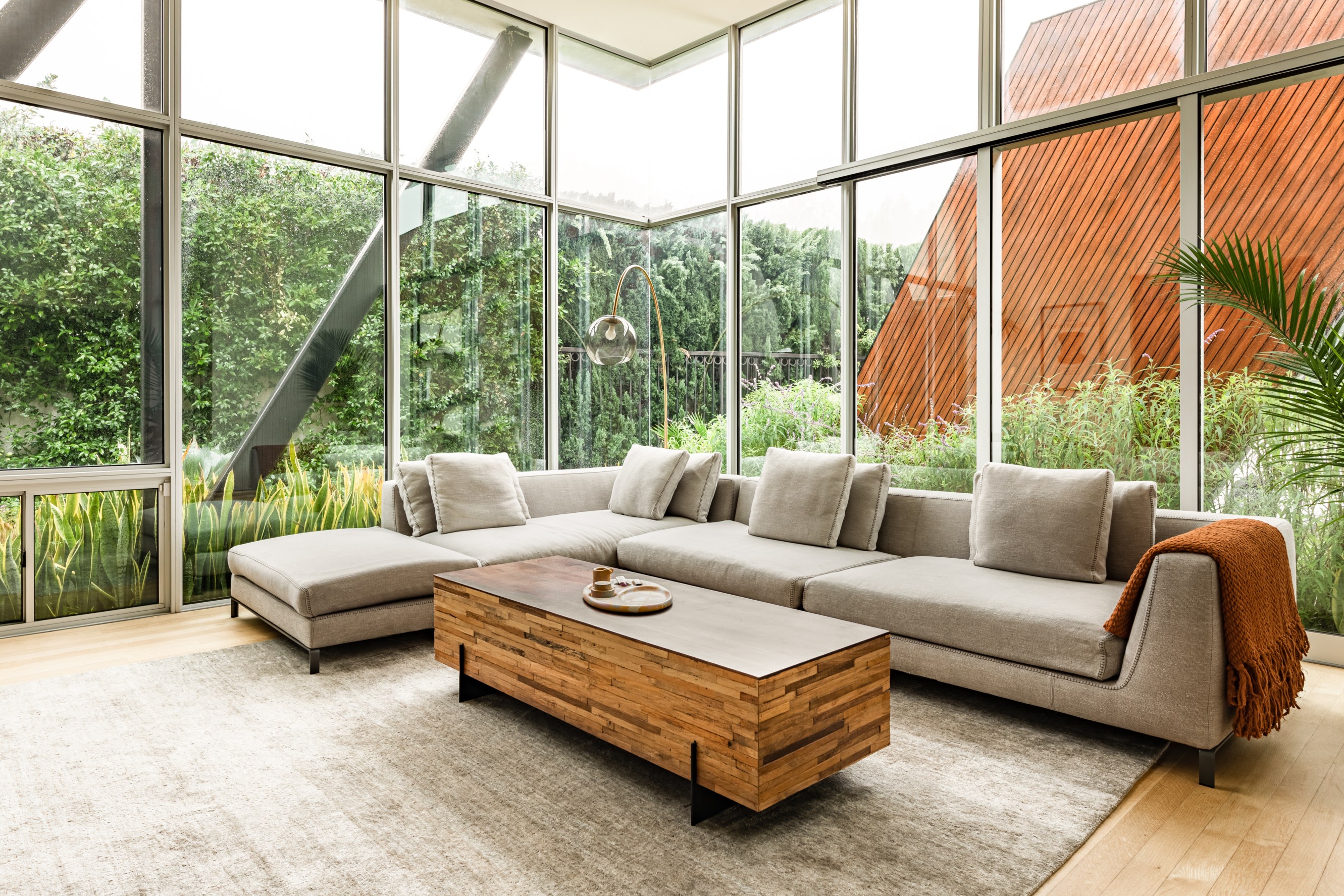 Houseplant home living room shot with floor to ceiling windows and a gray sectional couch and coffee table. Outdoor view includes a greenery wall that surrounds the house.