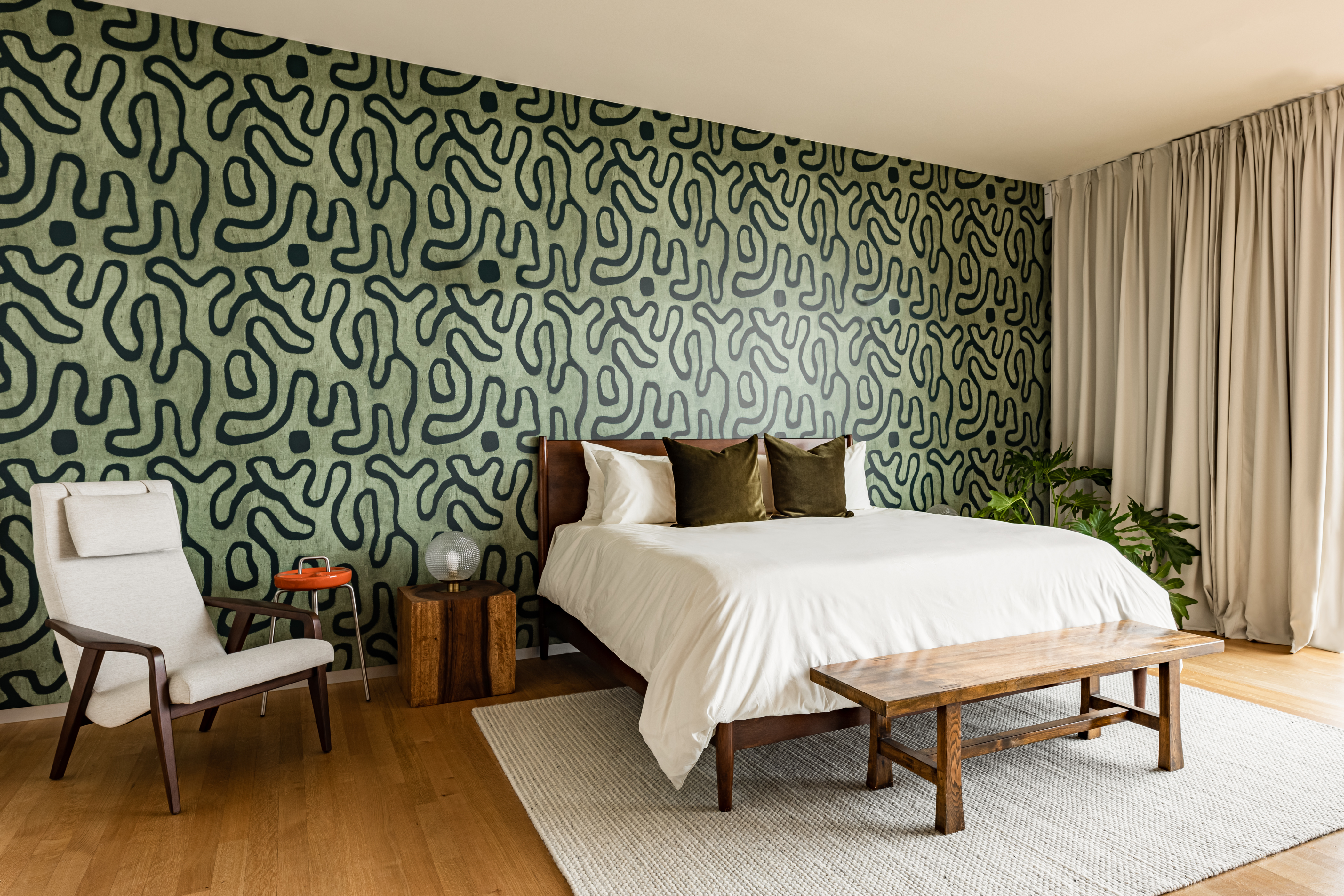 Houseplant home bedroom, which includes a large bed with white linens, a bench and side chair, with green wallpaper with abstract black designs on it.