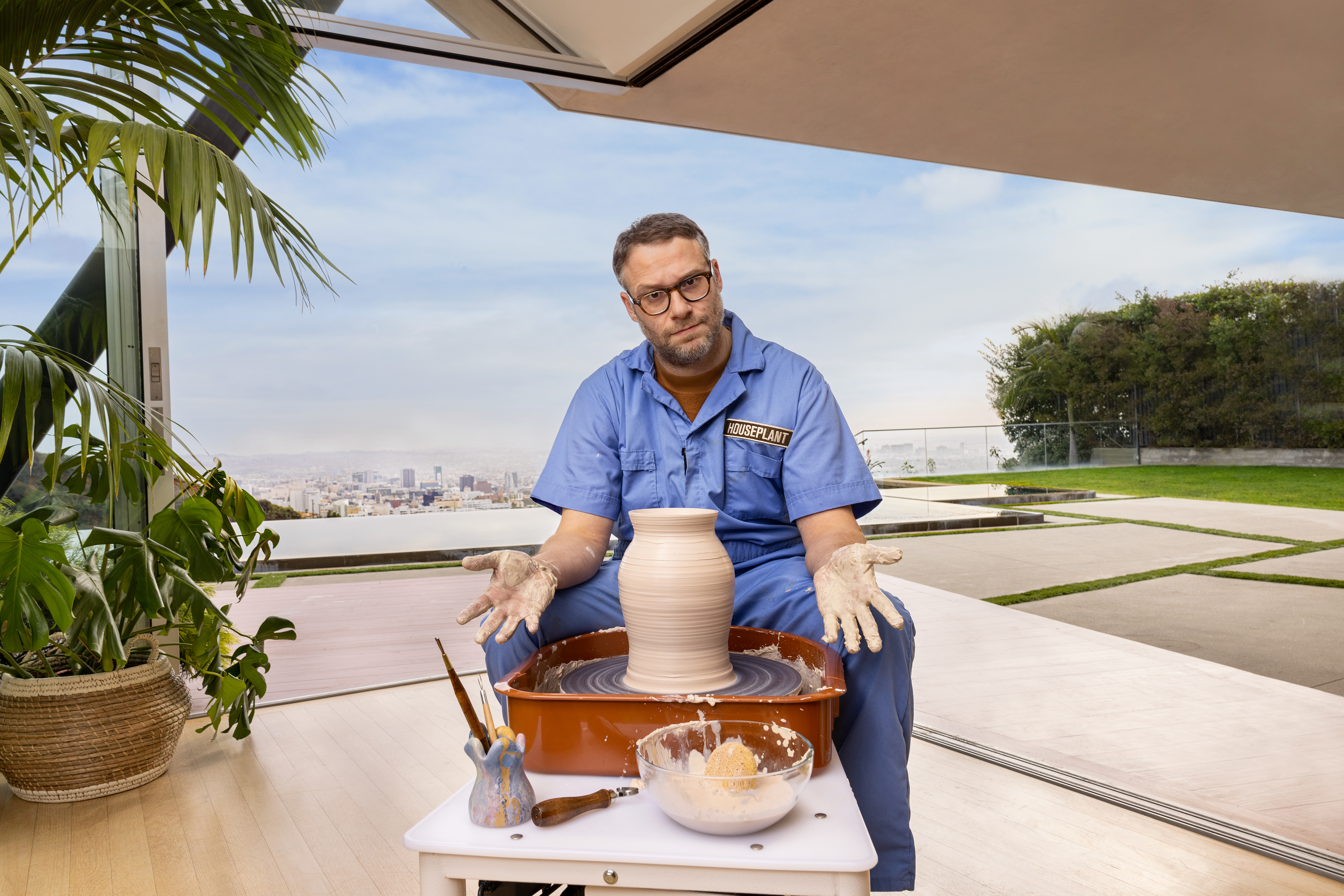 Seth Rogen in a blue art smock jumpsuit sitting at a pottery stool with a shrugging expression and a view of downtown Los Angeles in the background.