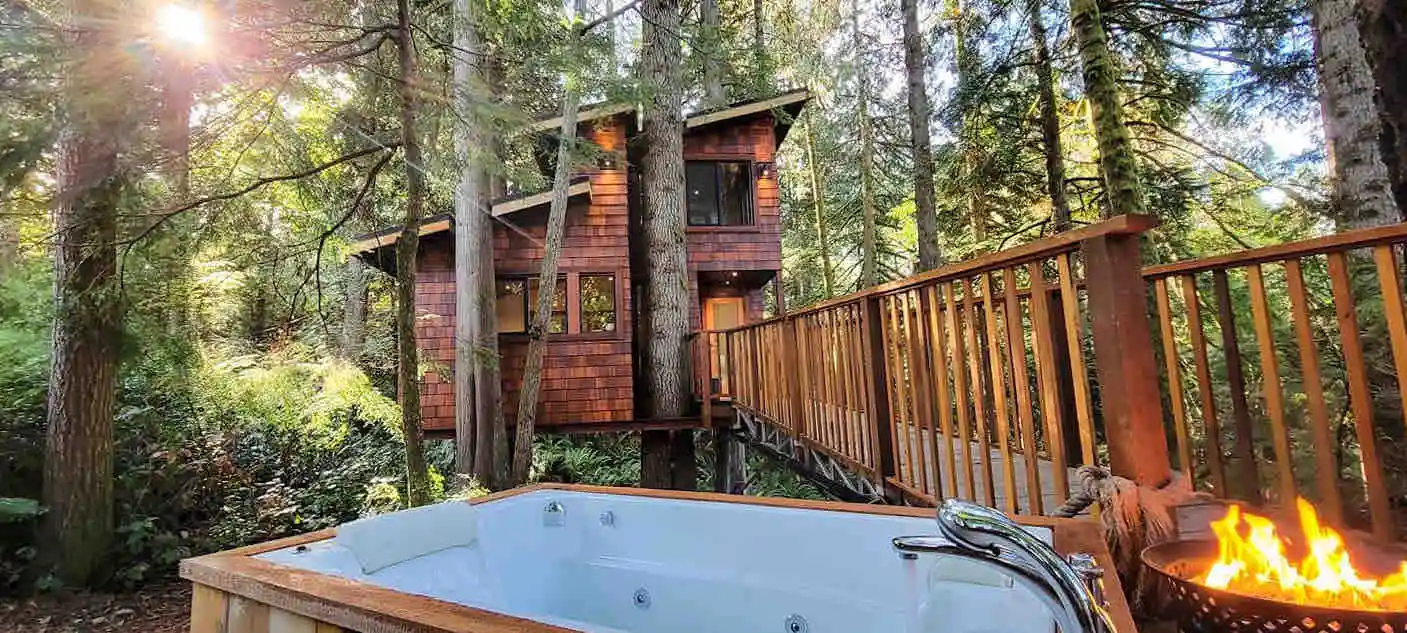 The sun peeks through the trees behind a wooden cabin with a large wooden walkway leading to an outdoor bath in the foreground. 