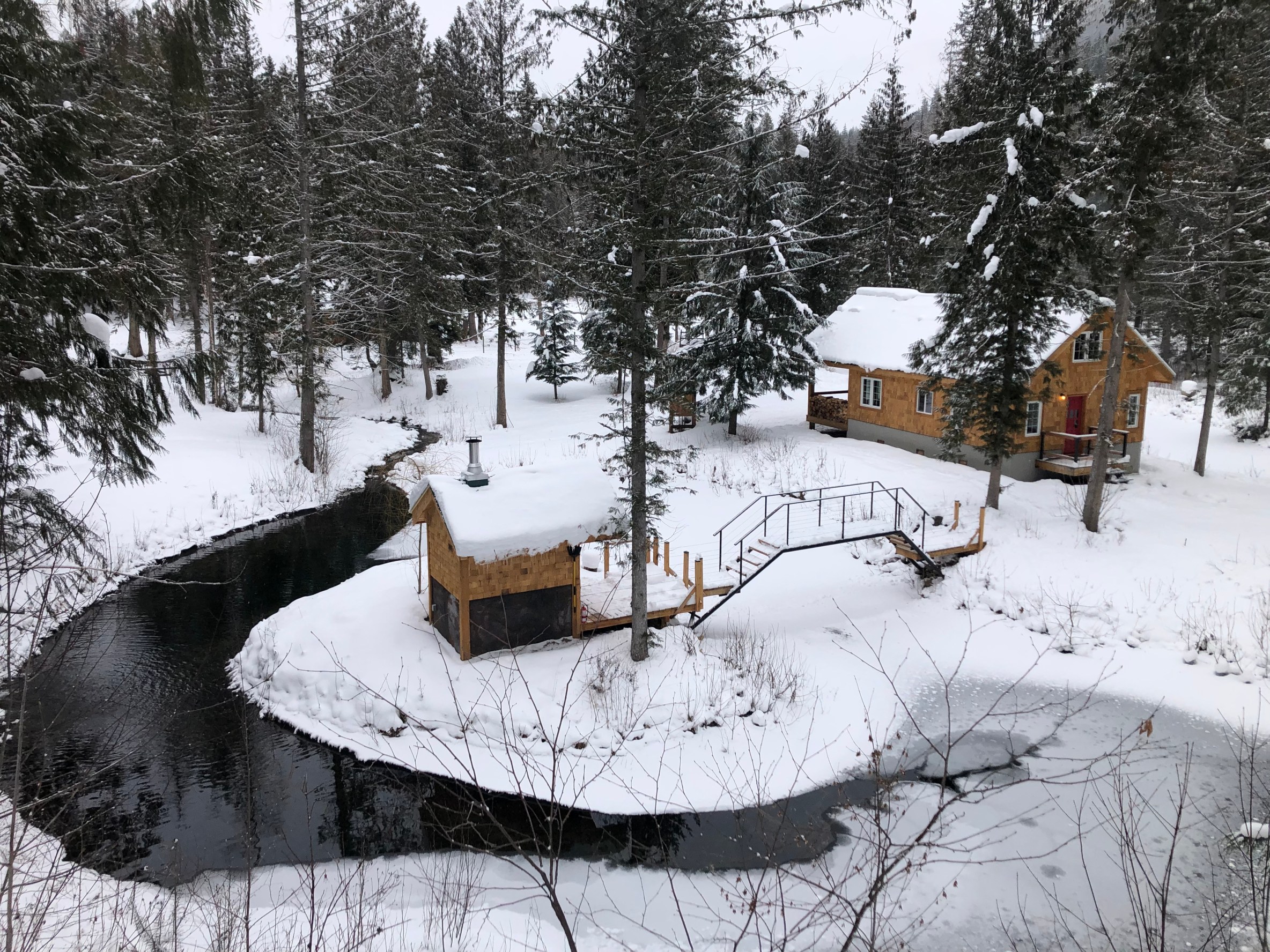 A snow capped sauna is surrounded by an icy stream and wintry trees in the foreground, with a walking bridge leading to a cabin behind it. 