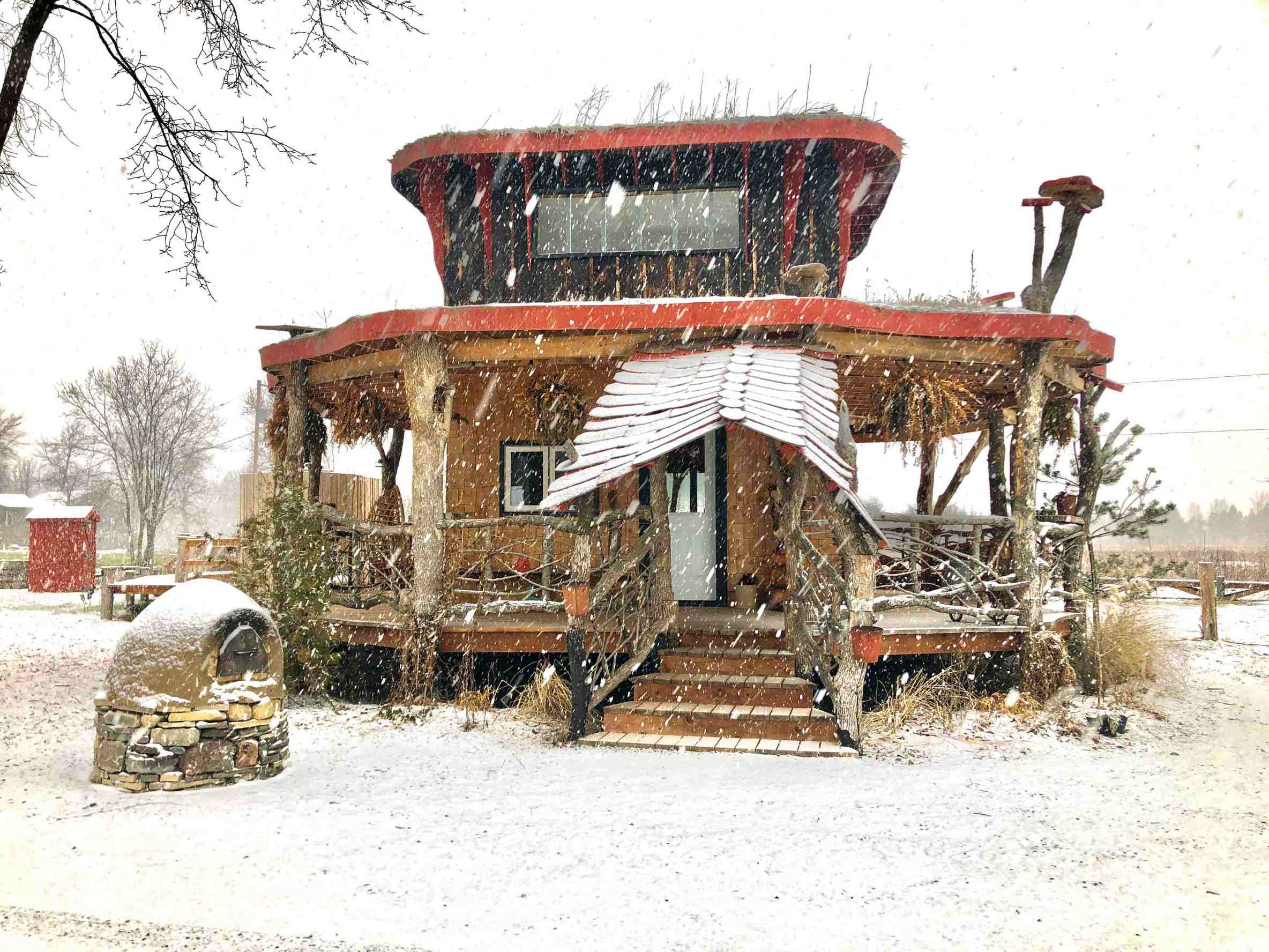 Snow is coming down on a unique wooden house, capped with mushrooms statues. 
