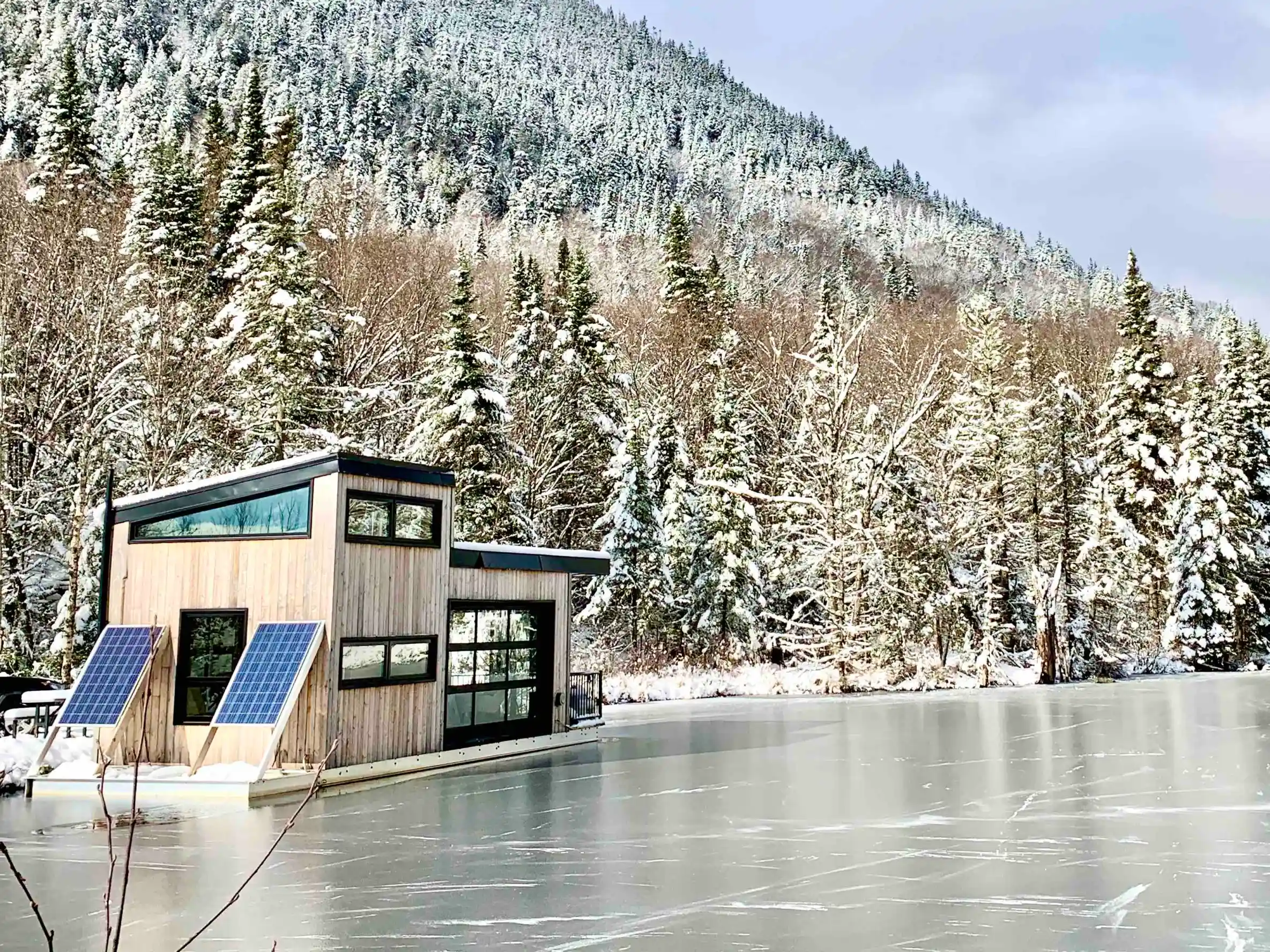 A modern boat chalet sitting on a lake frozen over. Behind it, a mountain with a forest of snow capped trees. 