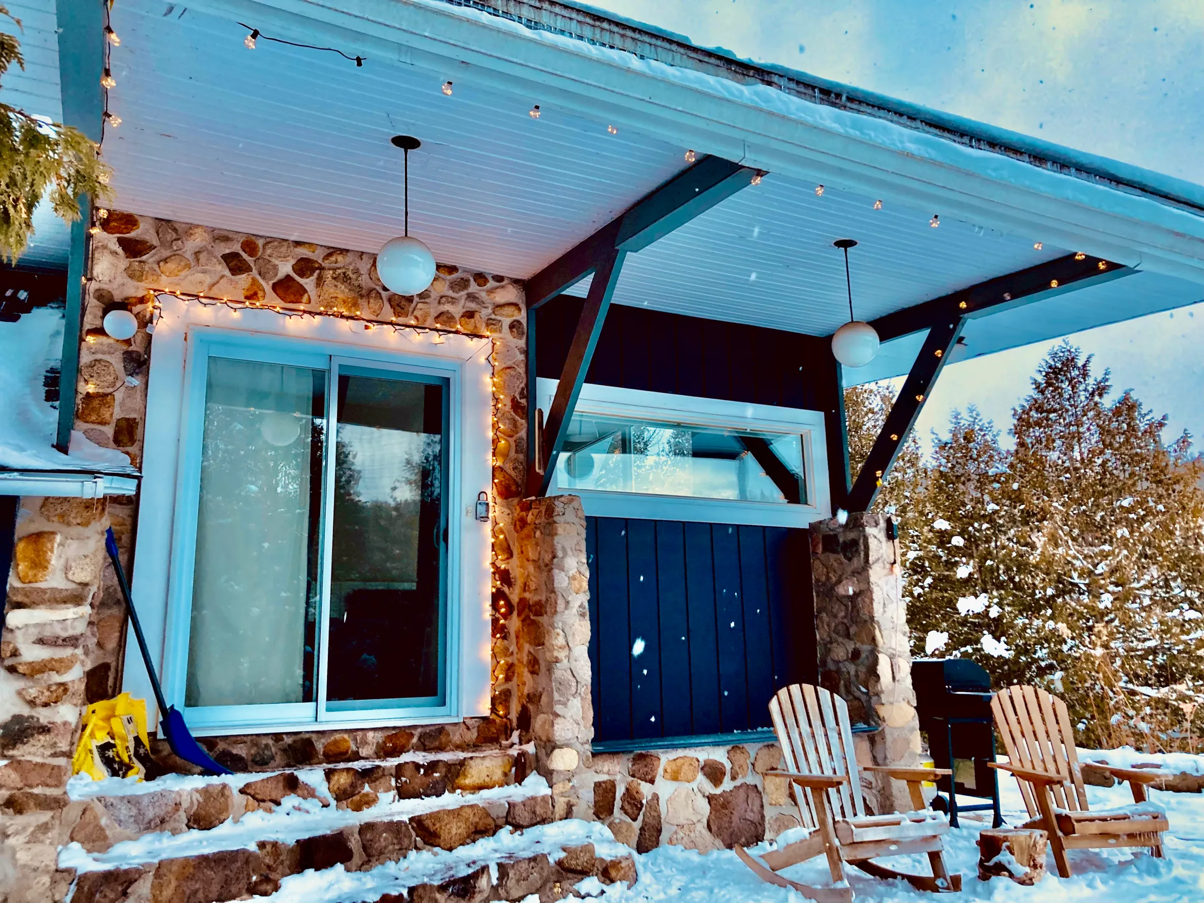 The side door view of a chalet as the snow falls. Two Adirondack chairs sit next to stony steps leading up to a sliding glass door.  
