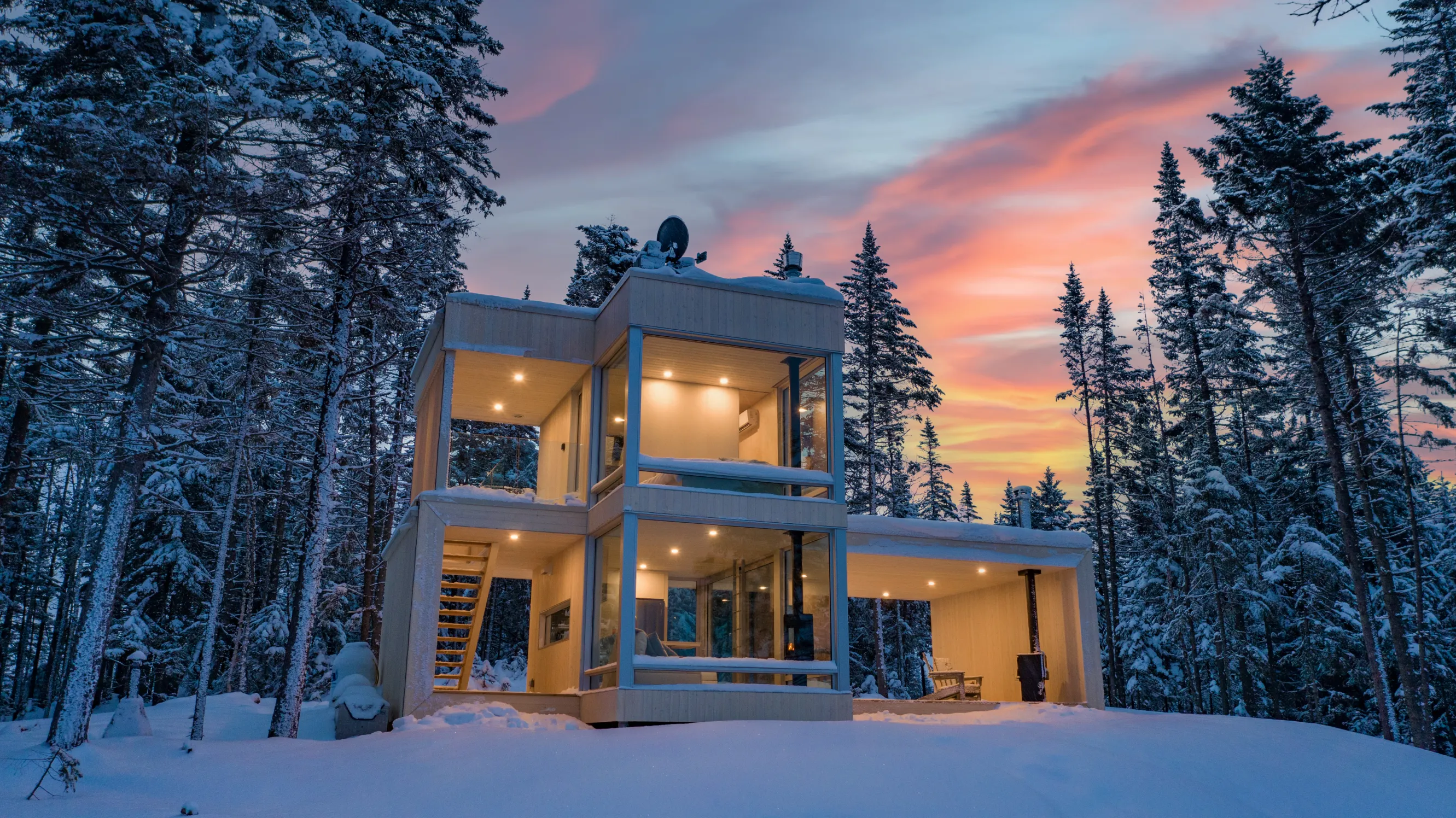 A modern two-story winter getaway atop a snowy hill surrounded by snowy trees, backed by a pink and yellow sunset sky.