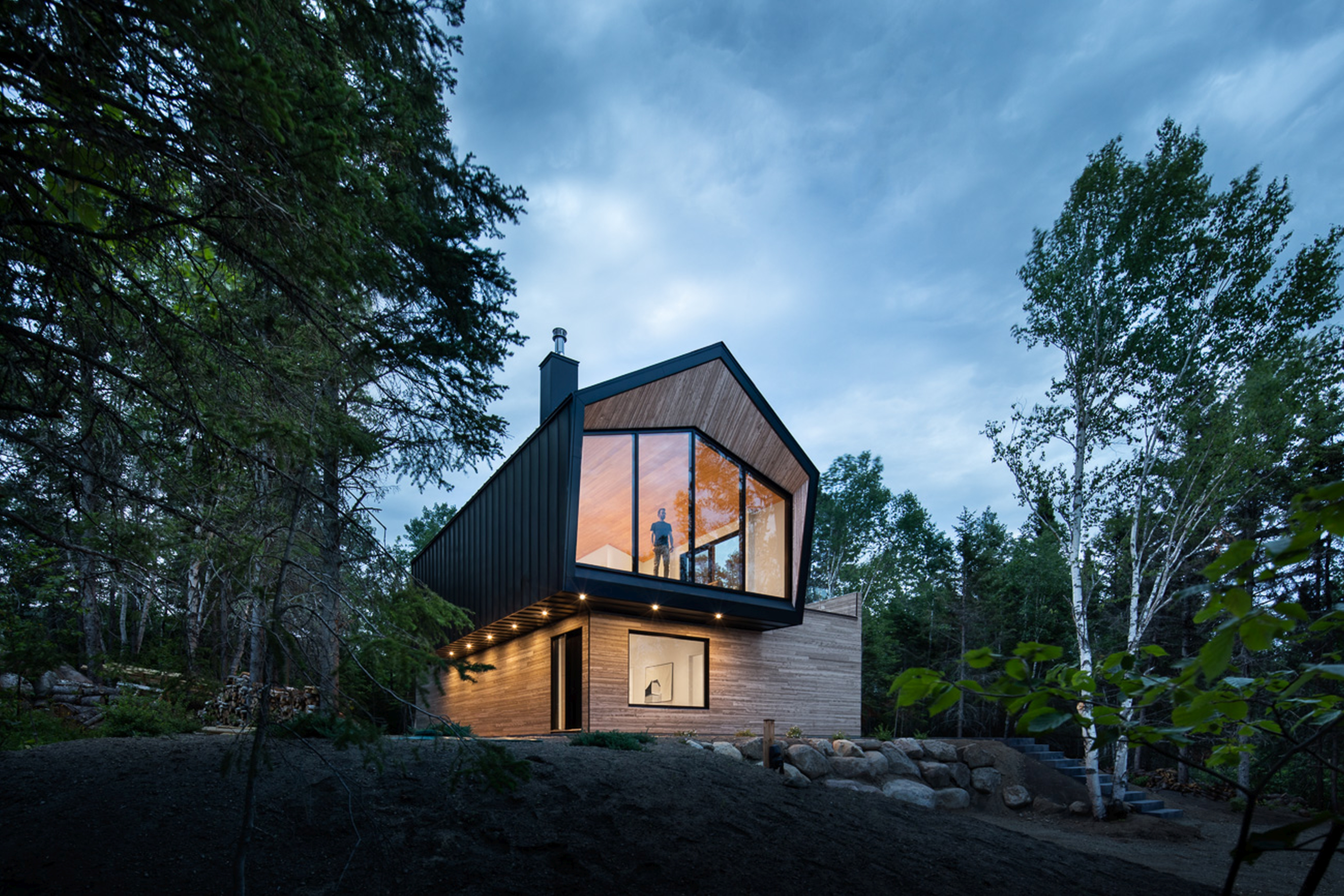 A modern and luxurious chalet at dusk with a person looking out of the floor to ceiling windows on the second floor.