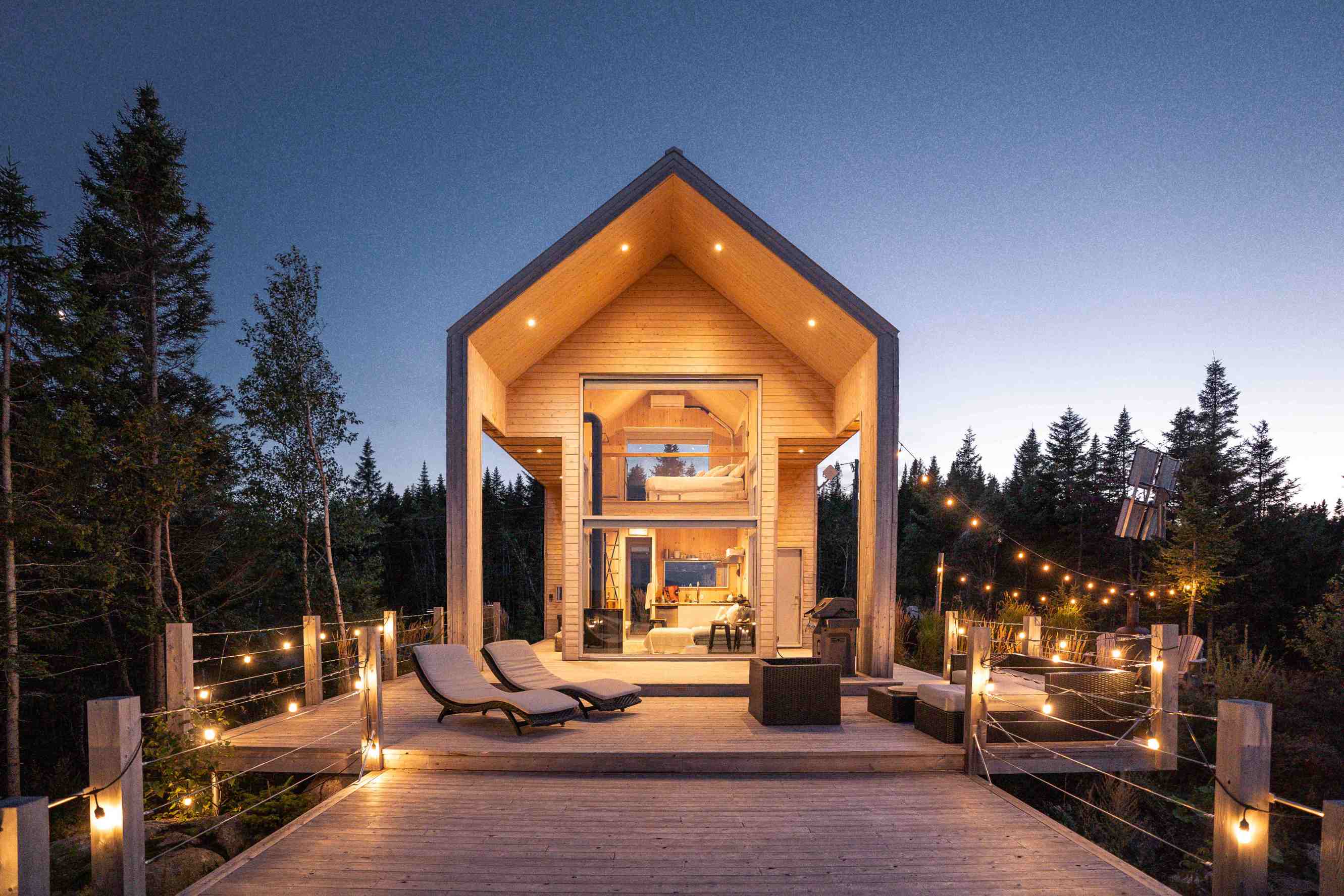 A modern two-story chalet at night with a large wooden patio at the entrance, lit by strings of outdoor lighting.