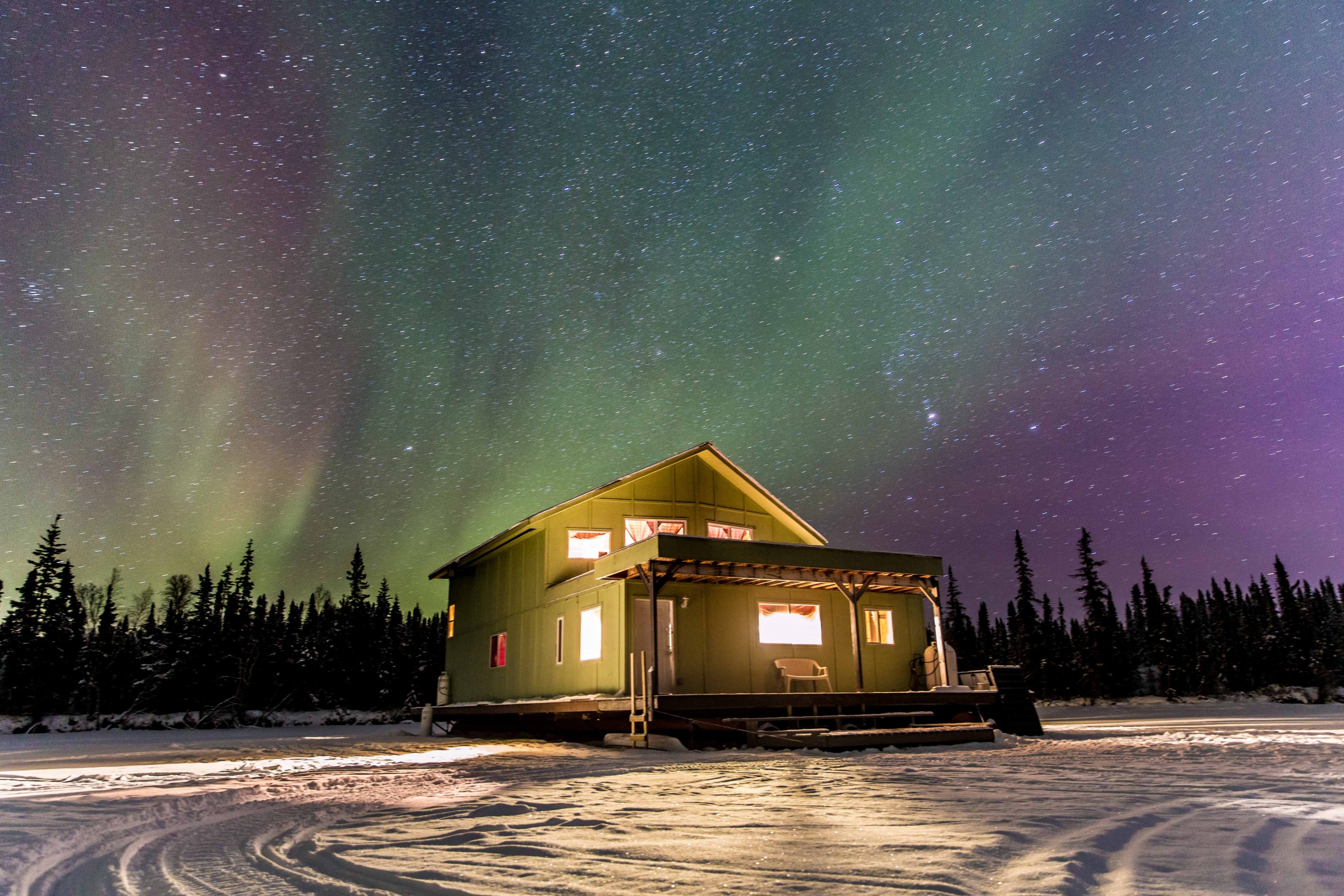 The northern lights dazzle the sky behind a green cabin sitting on in a flat, snowy field. 