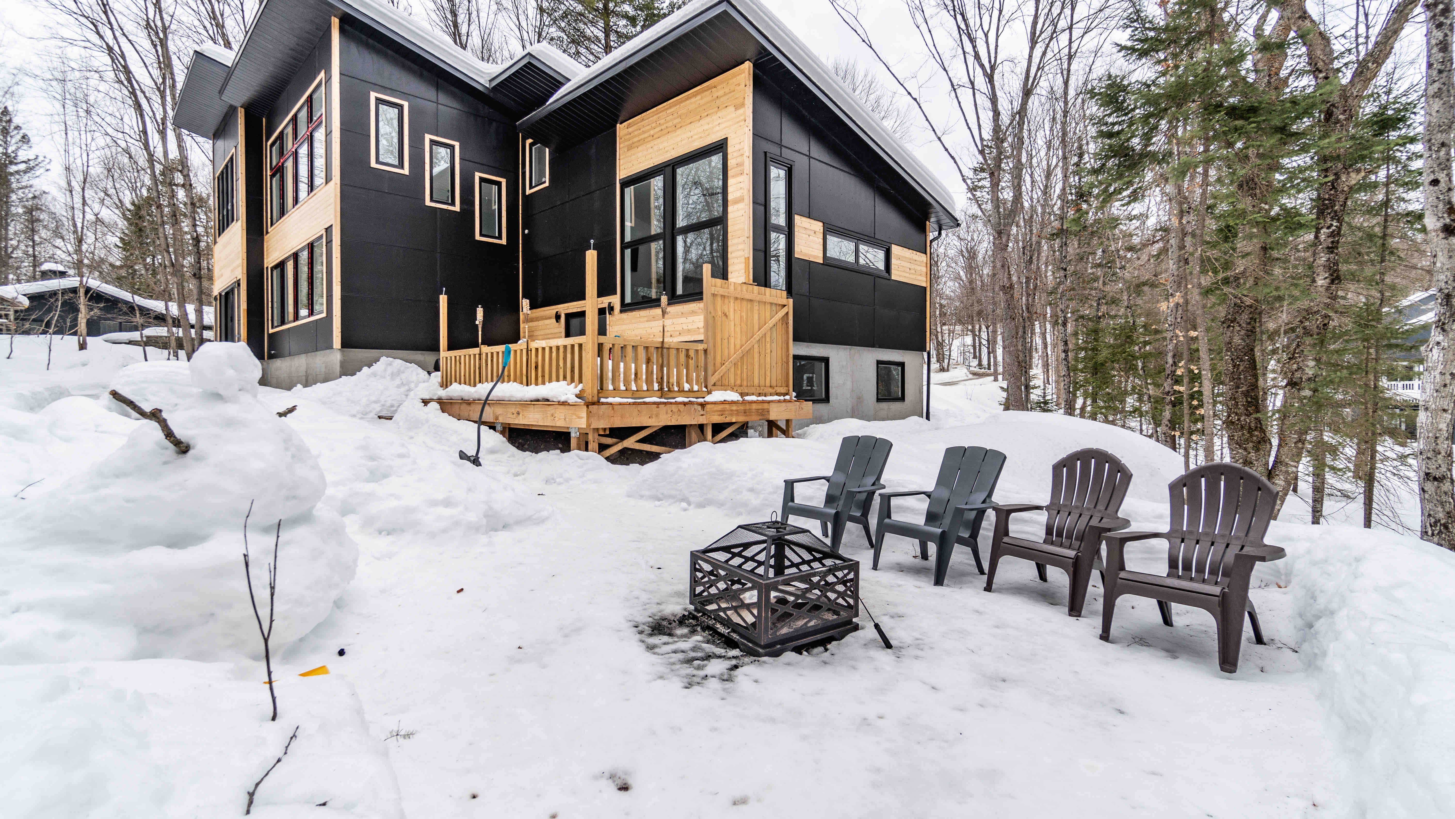 Four outdoor chairs and a firepit are sitting atop snow, with a modern multistory home behind. 