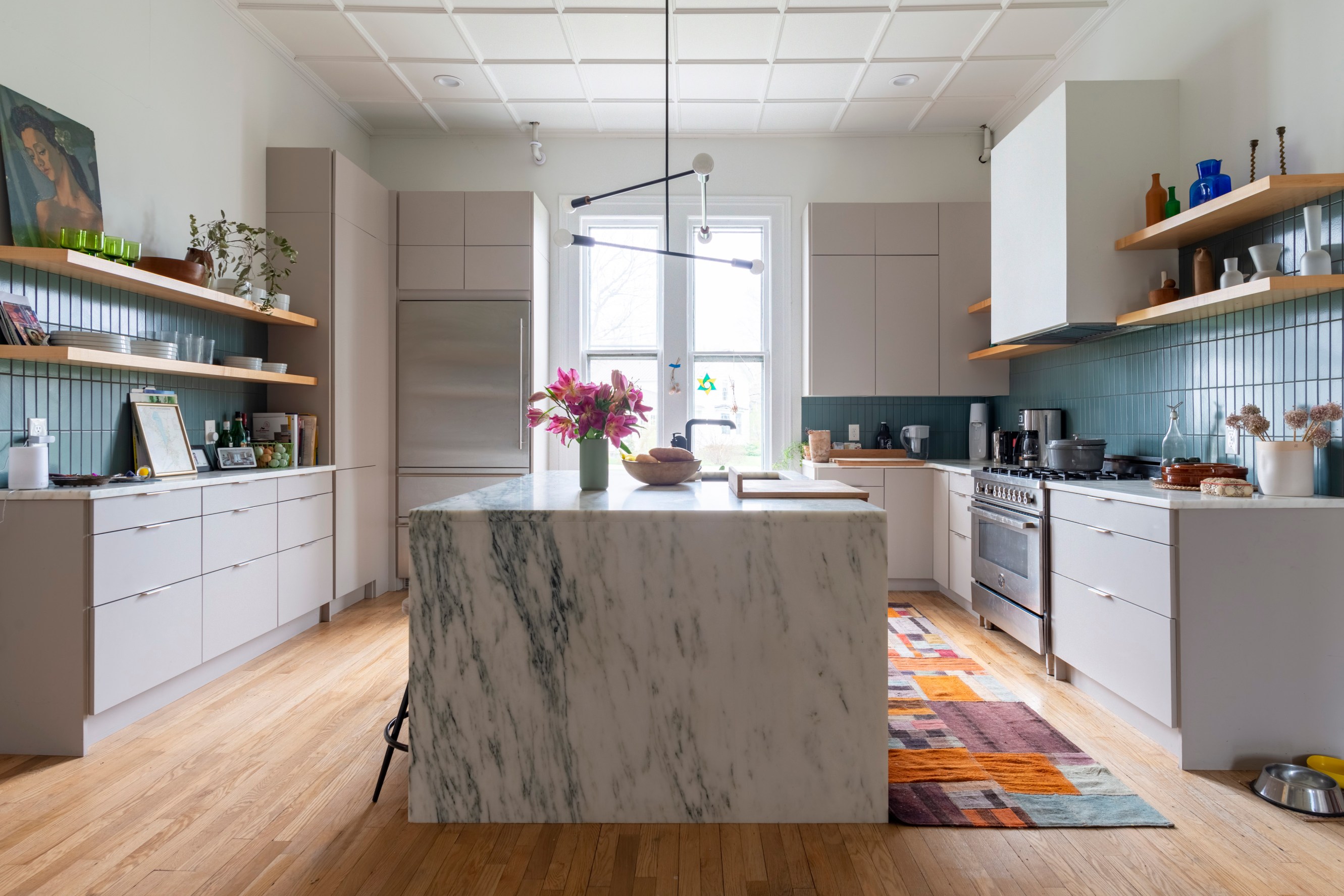 Bright and airy kitchen, with a white marble or quartz island and an industrial light fixture above it. Island is flanked by white countertops with robin's egg tile backsplash.