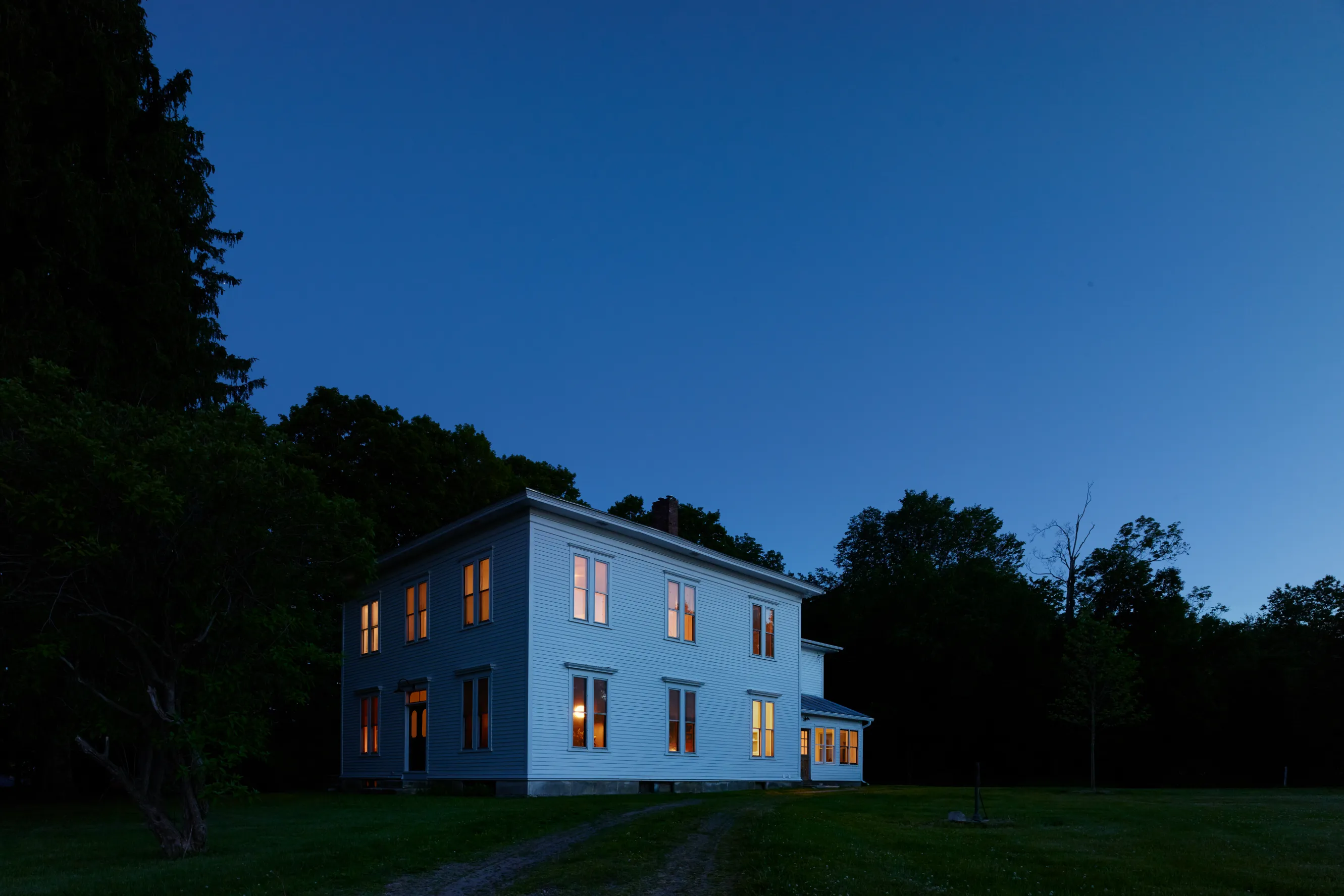 A night shot of a clear blue sky and a white two-story home in the center, surrounded by trees.