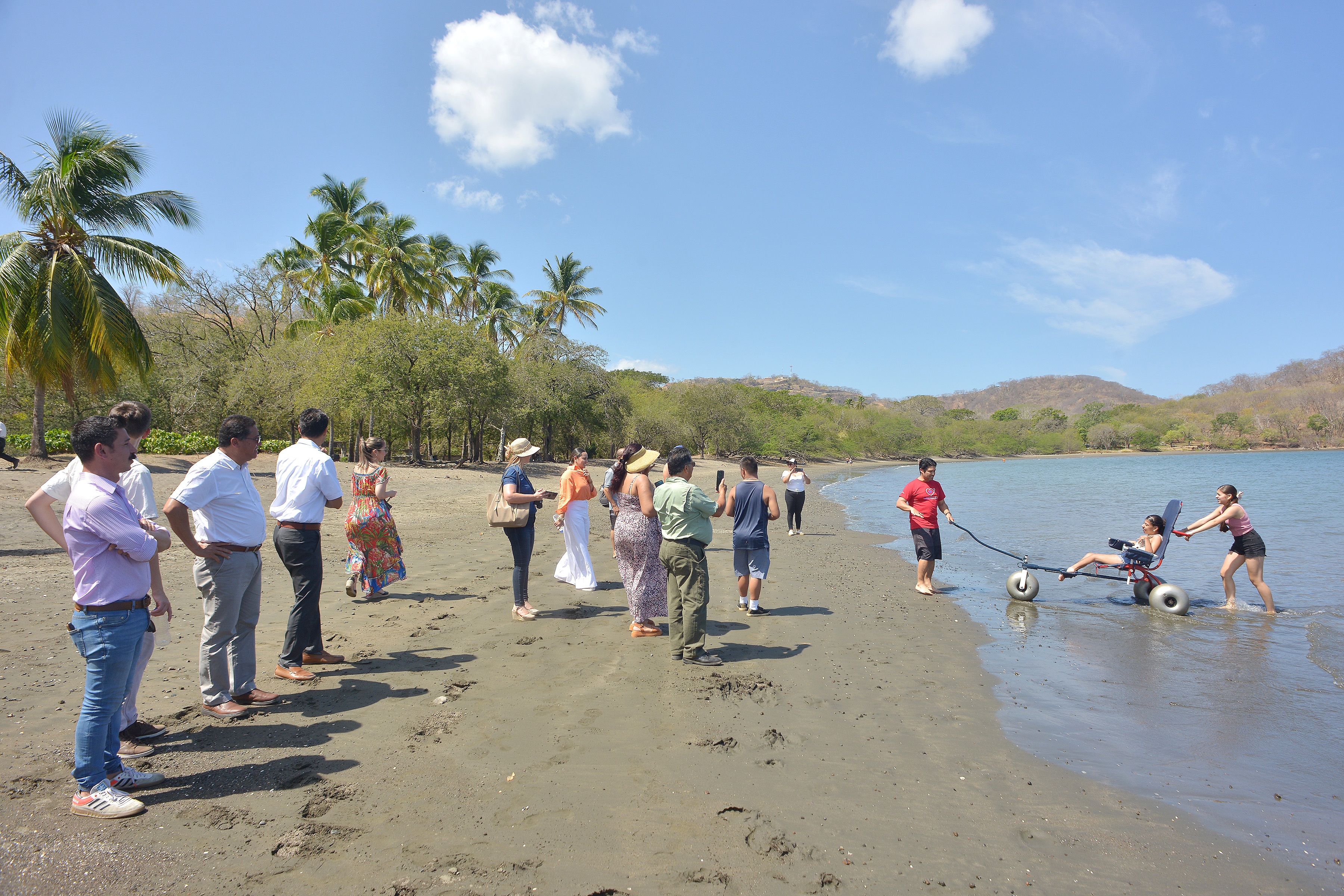Personas a la orilla de la playa de Costa Rica participando en la inauguración de la playa accesible. Una persona en silla de ruedas está dentro del mar con su cuidador y el equipamento requerido para disfrutar del mar