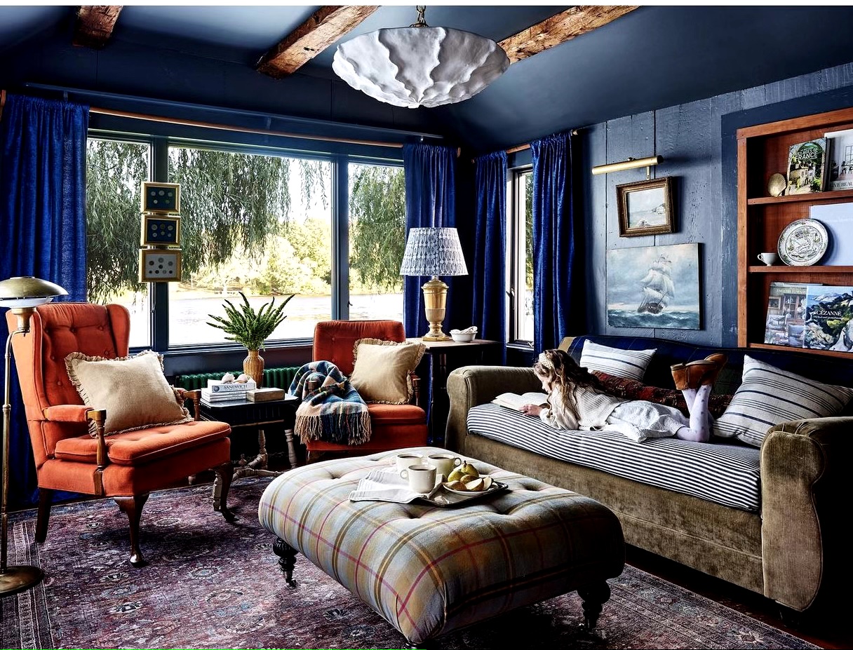 A blue-painted living room with a velvet sofa and two orange armchairs with a plaid footrest and antique art fixtures on the walls.