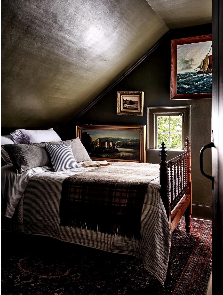 A moody, dark green bedroom with a wood frame bed with earthy colored linens and antique nautical and landscape oil paintings on the wall.