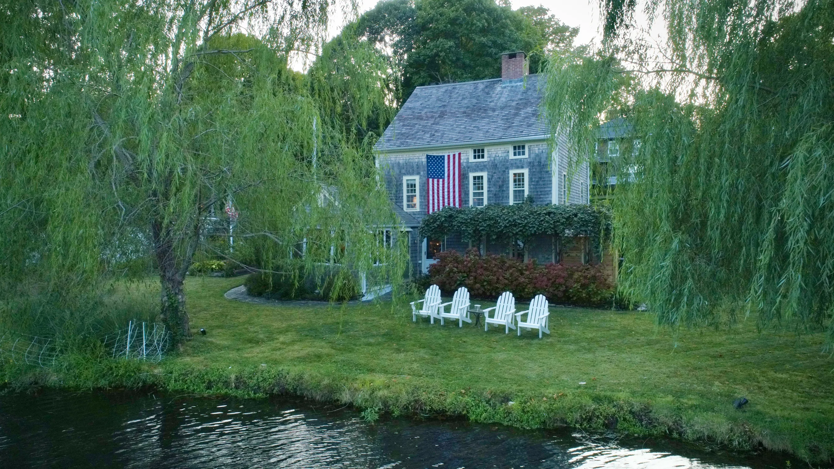 A backyard exterior shot of the home, showing a pond in the foreground with a backyard and four white Adirondack chairs with the house in the background, donning an American flag hanging from the second floor.