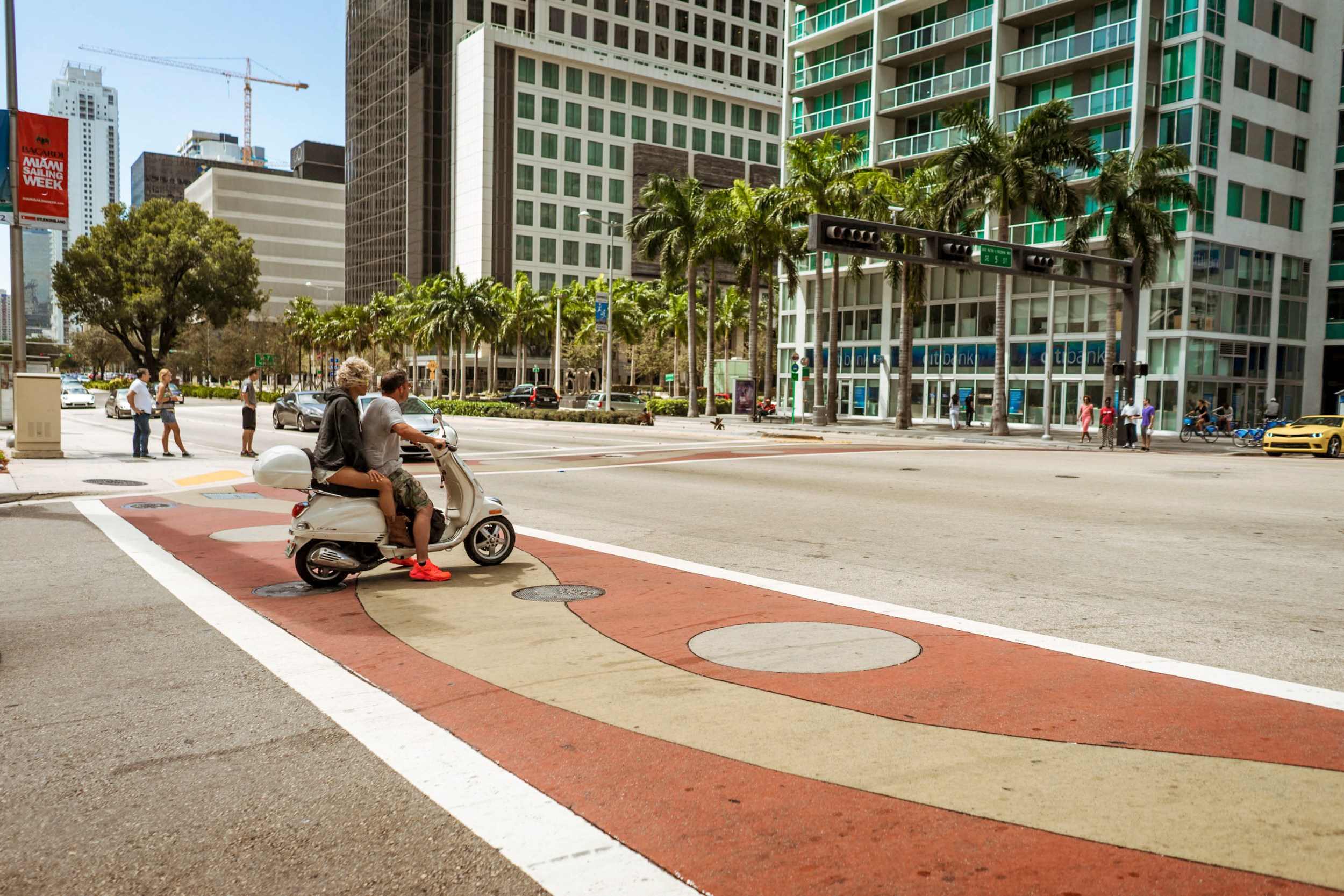 A bustling intersection in a downtown area with pedestrians, cars, and two people riding a scooter