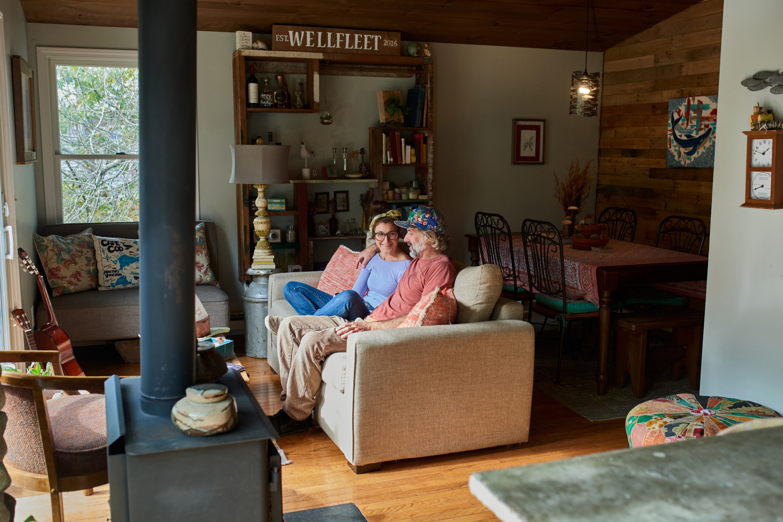 Man sits with his arm around a smiling woman on a couch in a quaint living room near a wood stove