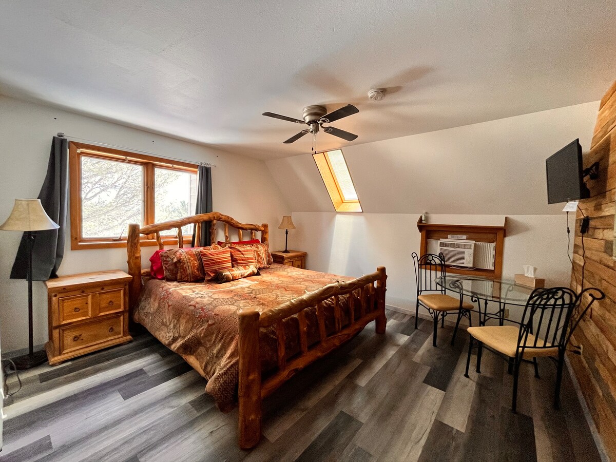 A bedroom with a double bed, ceiling fan, tv and two sitting chairs with table. 