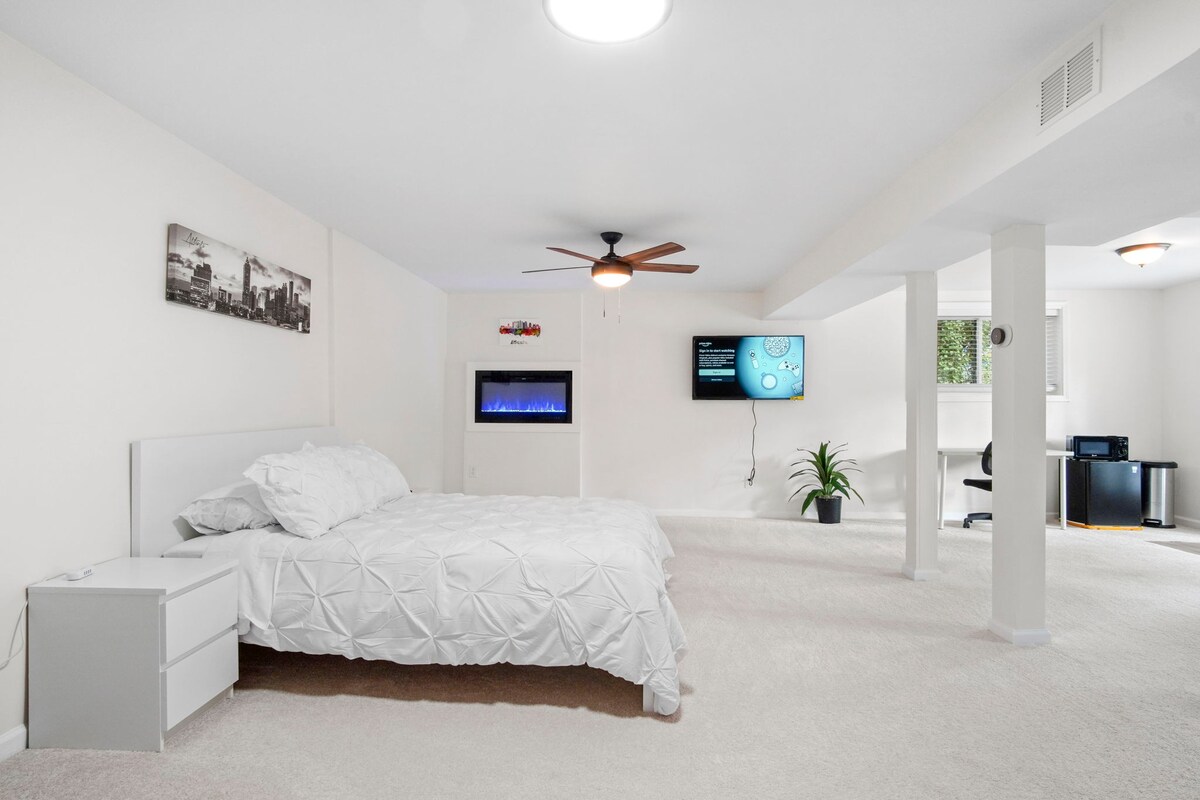A bedroom with white walls, white bed covers, tv on the wall and desk in the corner. 