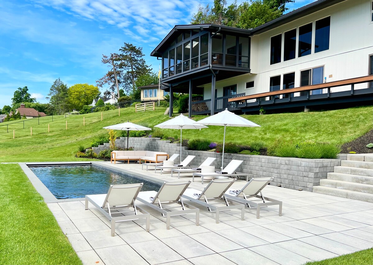 A pool surrounded by grey pavement and sun loungers with the house in the background. 