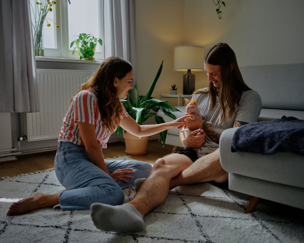 A woman in a striped shirt sits on the floor and pets the cat of a man in a gray shirt with long hair