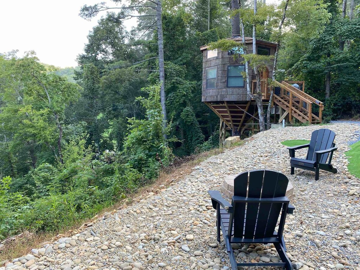 Treehouse with a forest with gravel driveway and two chairs in front of it. 