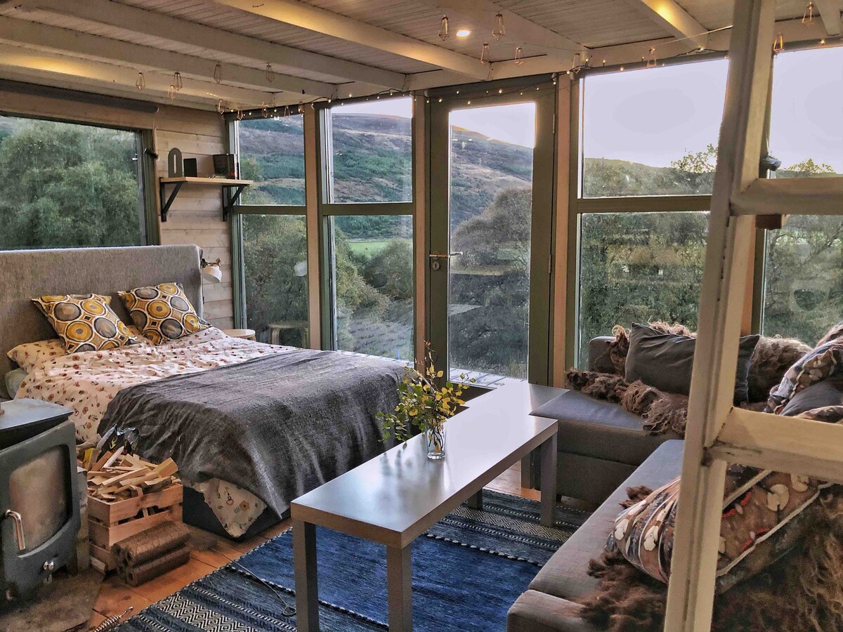 Picture of a double bed, sofa, and coffee table with floor to ceiling windows looking out to nature. 