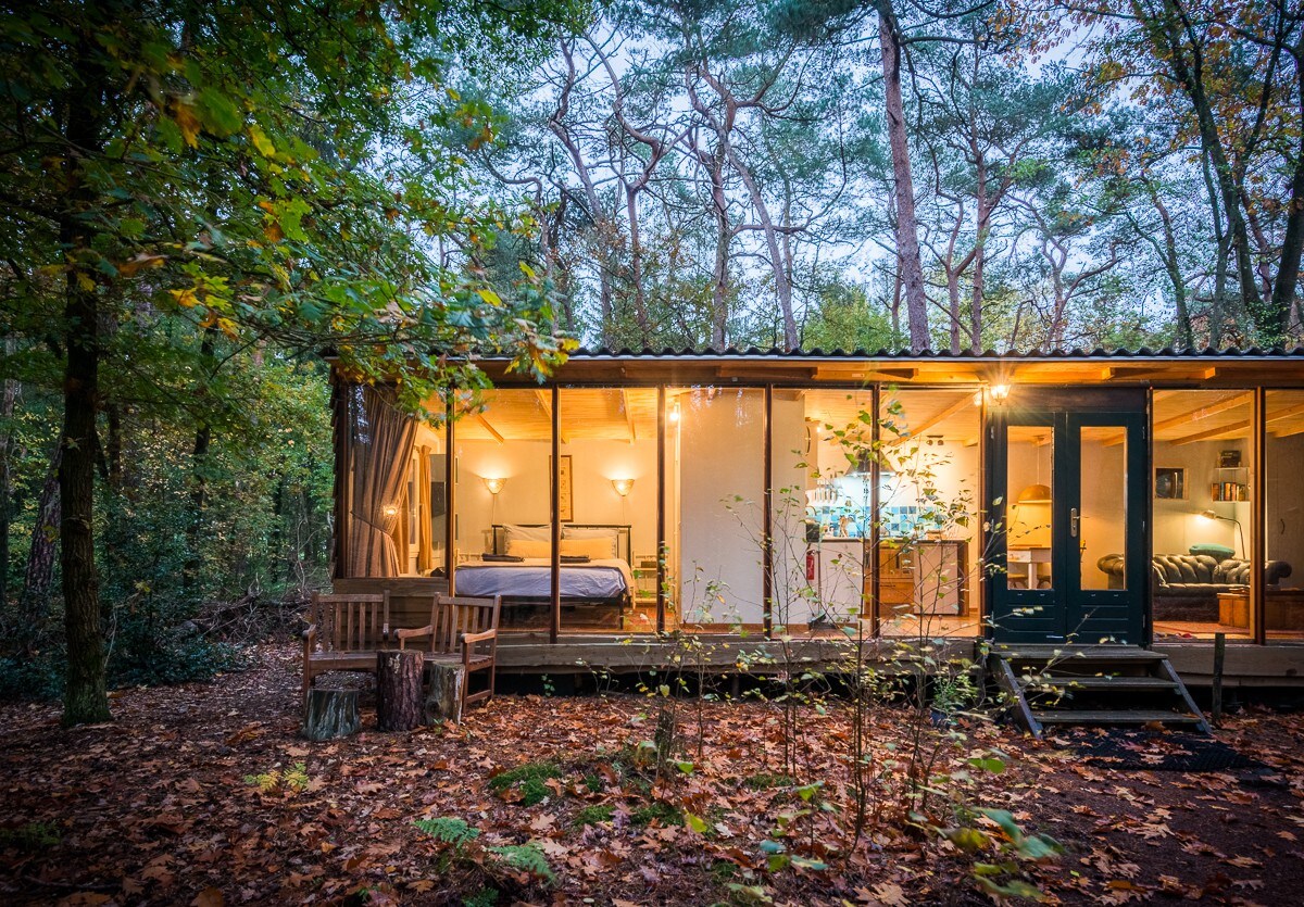 A picture of a low lit rectangular cabin in the wilderness with wooden chairs at the front of the cabin. 